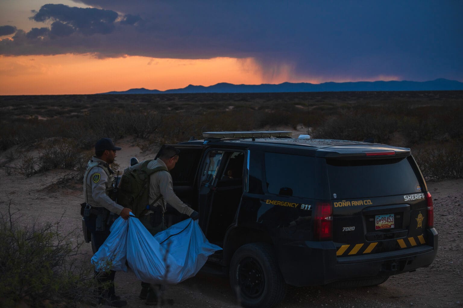 Two Doña Ana County sheriff’s deputies load a body bag into a patrol SUV at dusk in the desert, after recovering the remains of a migrant.
