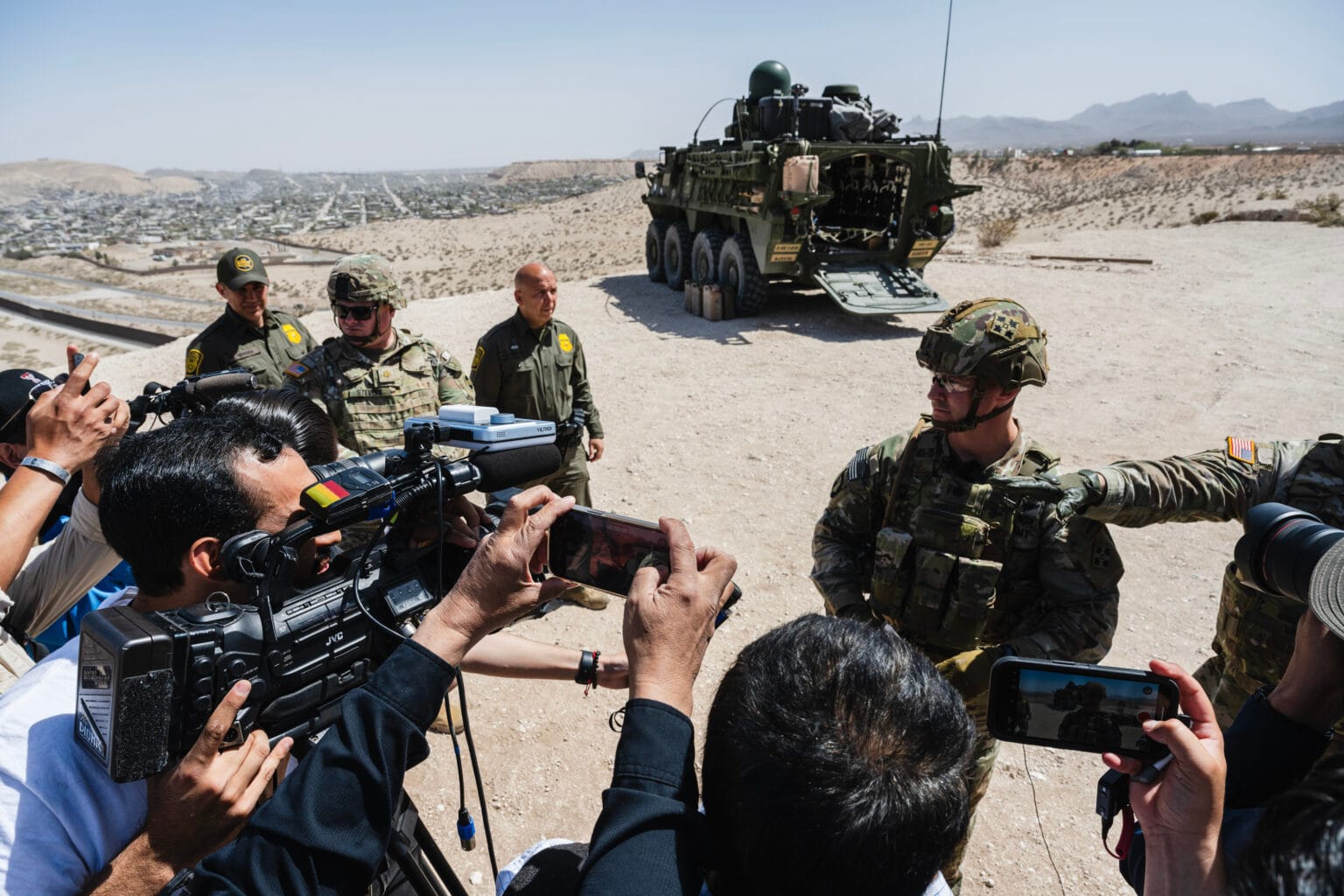 Members of the media surround U.S. military personnel during a press conference in Sunland Park, New Mexico, where CBP and Joint Task Force Southern Border announced the deployment of M1126 Infantry Carrier Vehicles to the El Paso sector on March 28, 2025. A military vehicle is visible in the background.