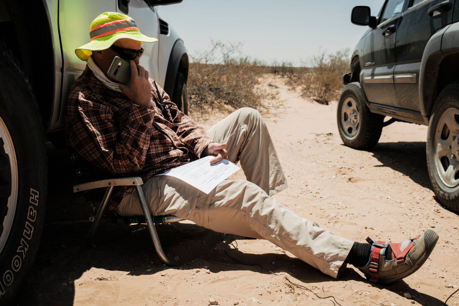 A man in a neon sunhat and plaid shirt sits in a folding chair between two vehicles in the desert, holding a cellphone to his ear and a document in his lap. Dry brush and sandy terrain stretch into the distance.