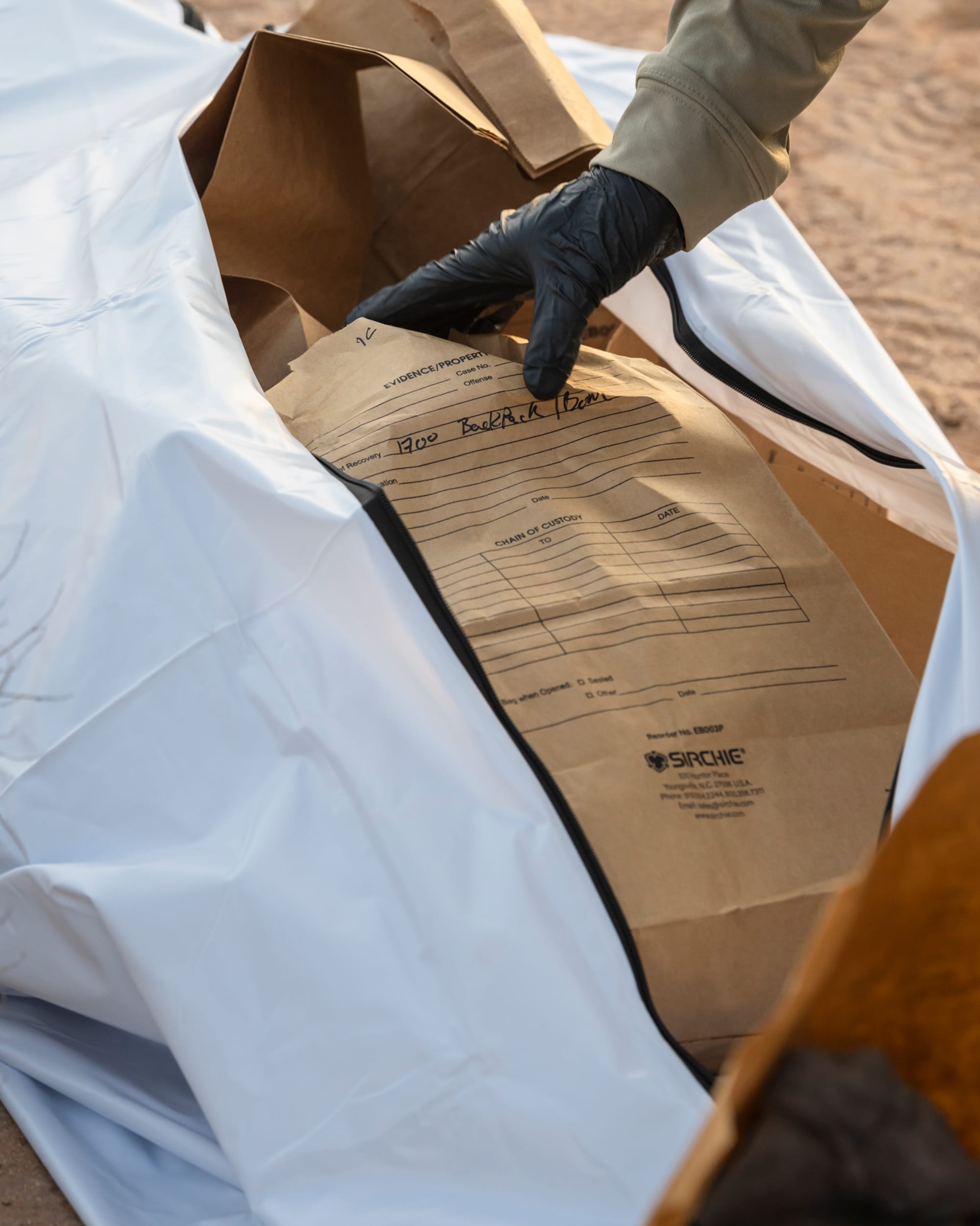 A gloved hand places a brown evidence bag labeled “Backpack (Bones)” into a white body bag during the collection of migrant remains in the desert.