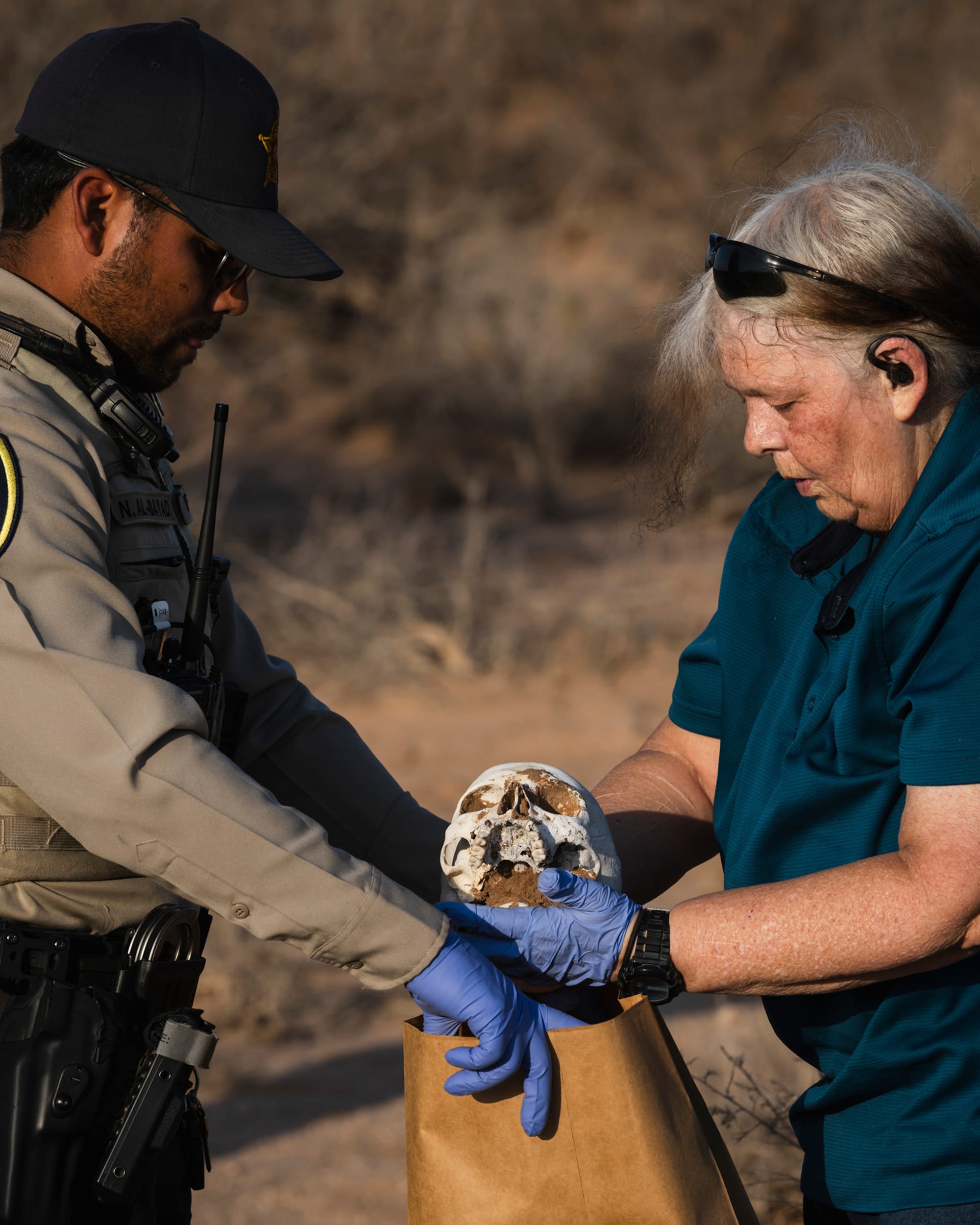 Doña Ana County Deputy N. Al-Abayad and field investigator Tambri Hunteman place a human skull into a brown evidence bag while wearing blue gloves in the desert.