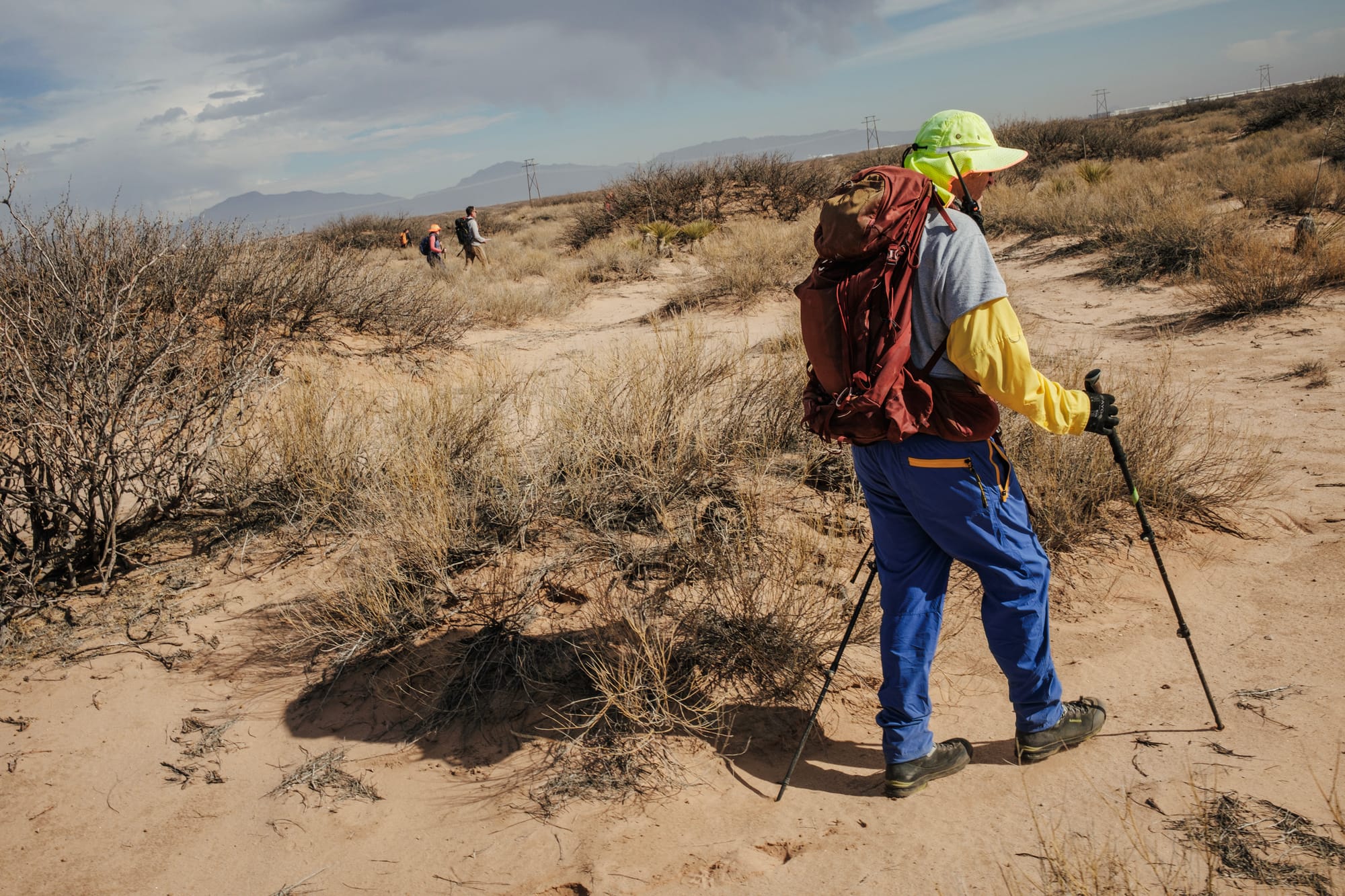 James Holeman, wearing a neon hat, hiking gear and carrying trekking poles, walks through desert brush while leading a volunteer search for migrant remains near the U.S.-Mexico border.