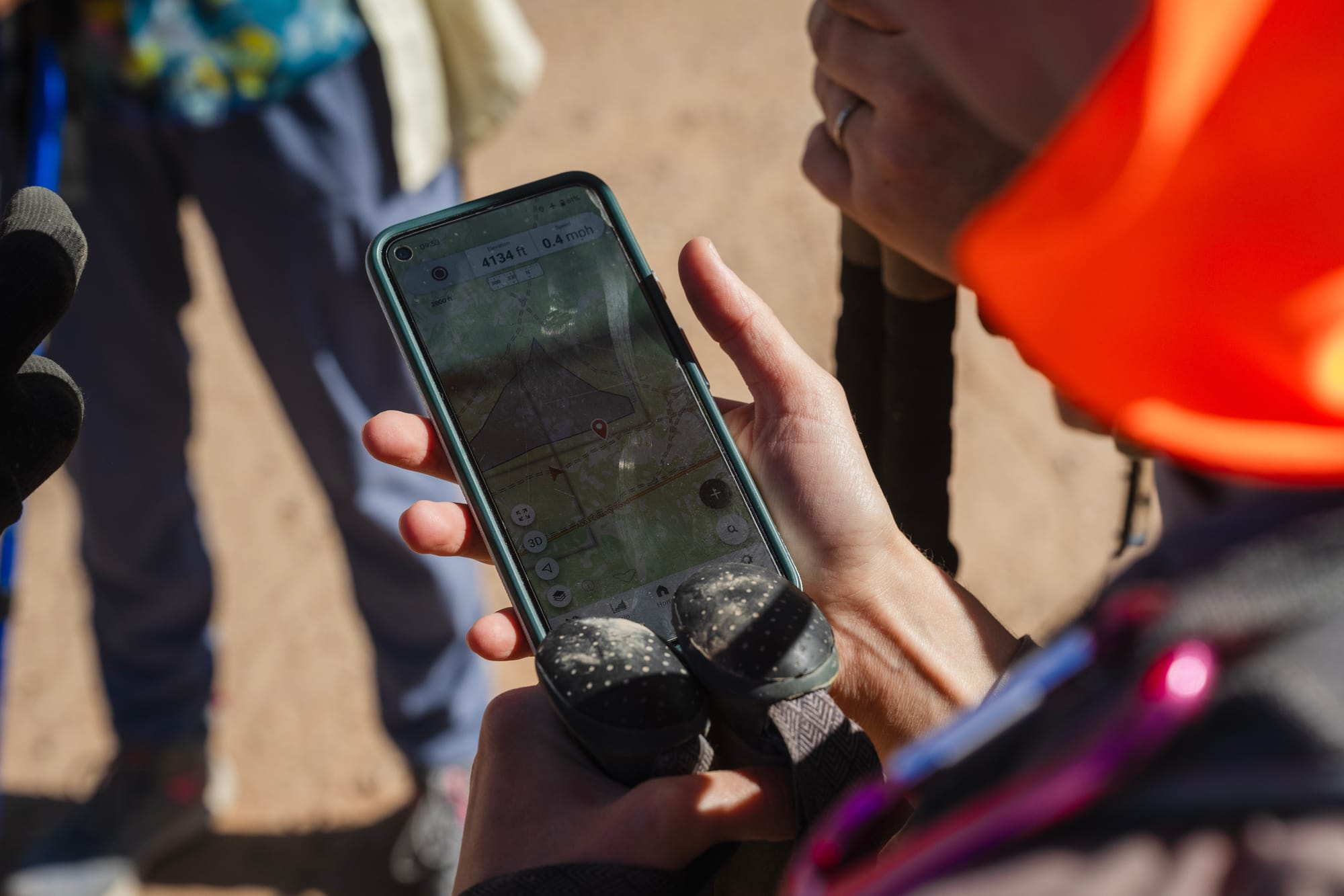 Close-up of a volunteer’s hand holding a smartphone with a map app open, showing coordinates during a desert search for migrant remains. Other volunteers and walking sticks are visible nearby.
