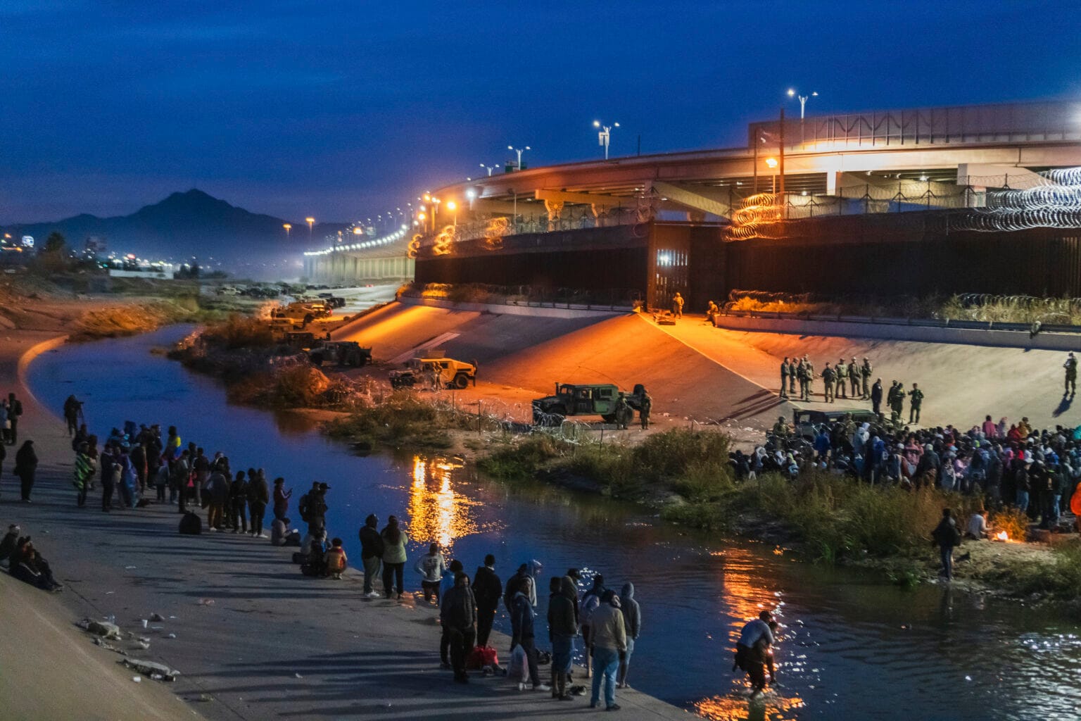 Migrants gather near the Rio Grande at night, facing rows of Texas National Guard troops and military vehicles blocking access to the border wall in El Paso. Floodlights and razor wire line the structure under a dark blue sky.