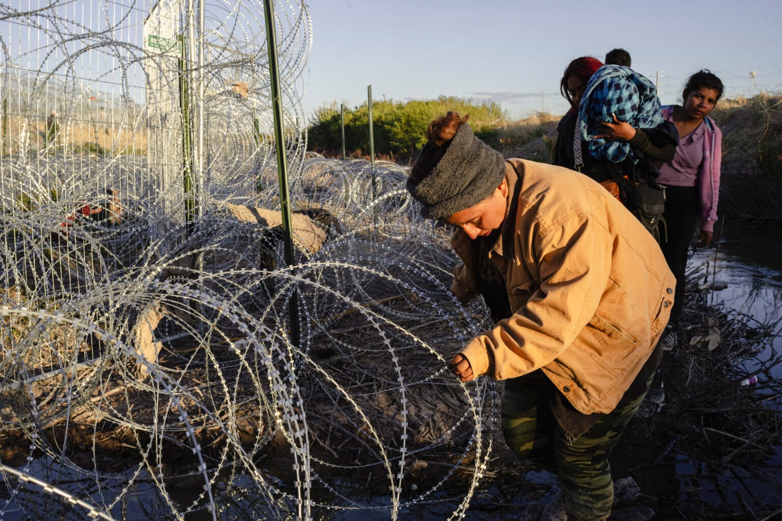 A woman carefully navigates through tangled concertina wire along the U.S.-Mexico border in El Paso at sunrise, while other migrants, including one carrying a child, wait behind her in muddy terrain.
