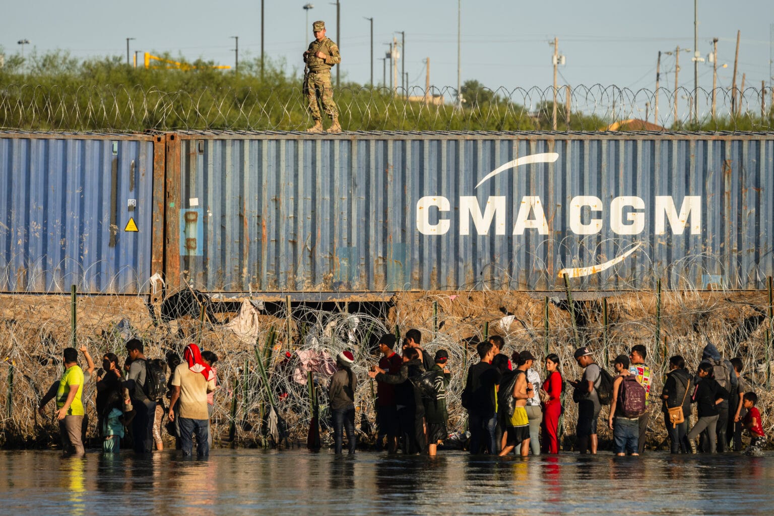 A group of migrants stands in shallow water at the U.S.-Mexico border in El Paso, facing a barrier of razor wire and shipping containers, as a Texas National Guard soldier watches from atop the container wall.