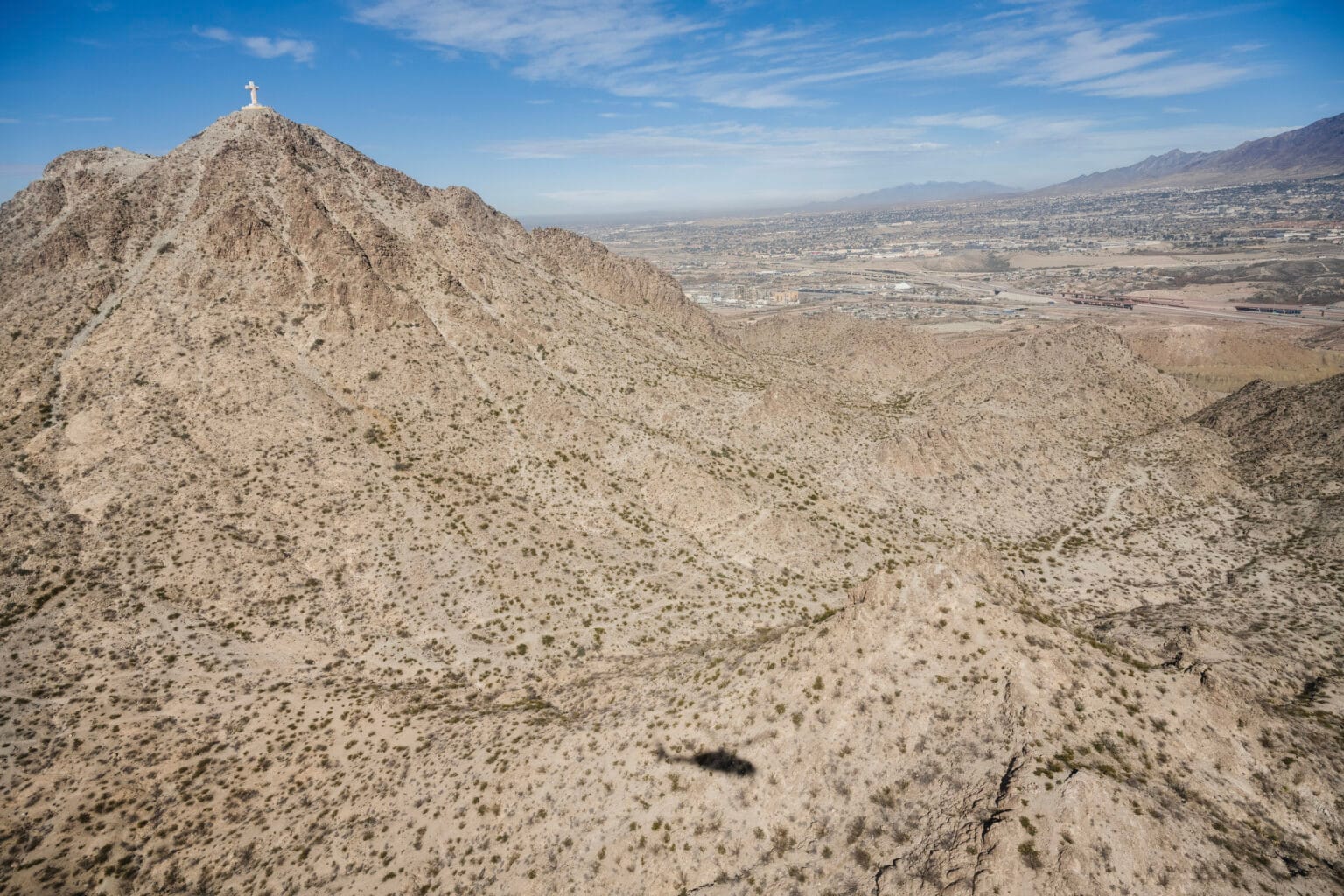 Aerial view of Mt. Cristo Rey in Sunland Park, New Mexico, with the shadow of a U.S. Customs and Border Protection helicopter visible over the rugged desert terrain. The mountain is a frequent crossing point for migrants and smugglers entering the El Paso sector.