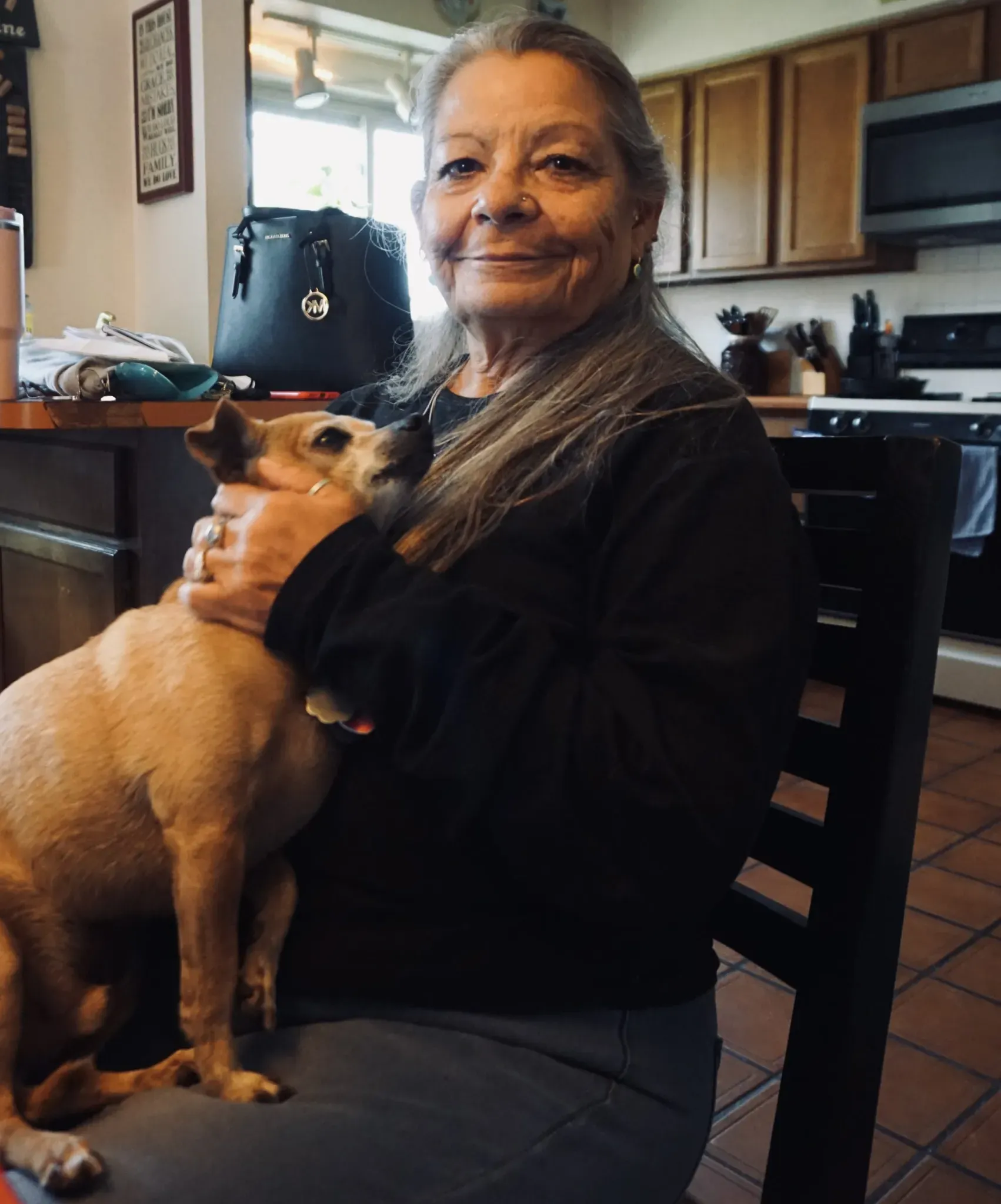 Older woman with long gray hair sitting in a kitchen, holding a small tan dog in her arms and smiling softly at the camera
