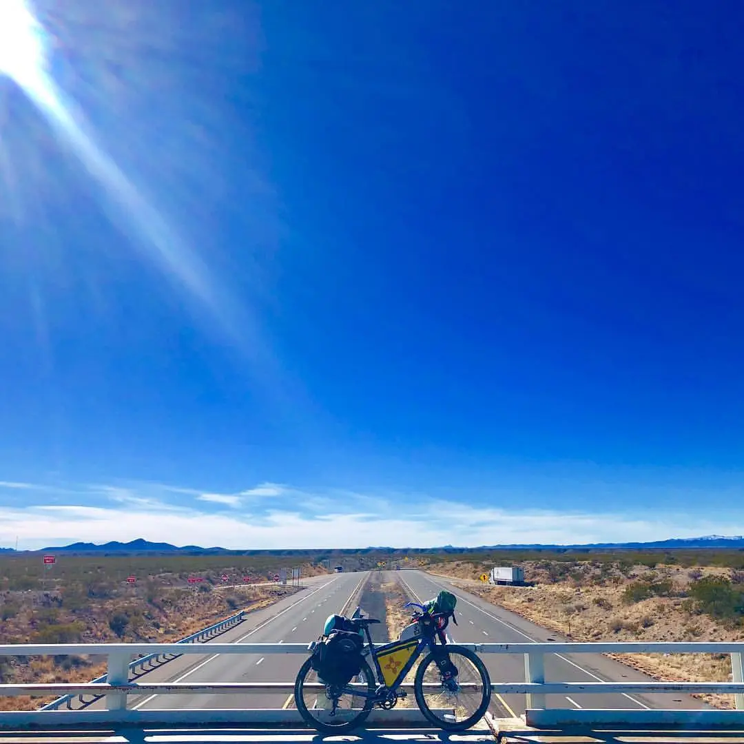 Bicycle loaded with gear parked on an overpass railing, overlooking a wide desert highway under a bright blue sky with the sun beaming from the upper left corner