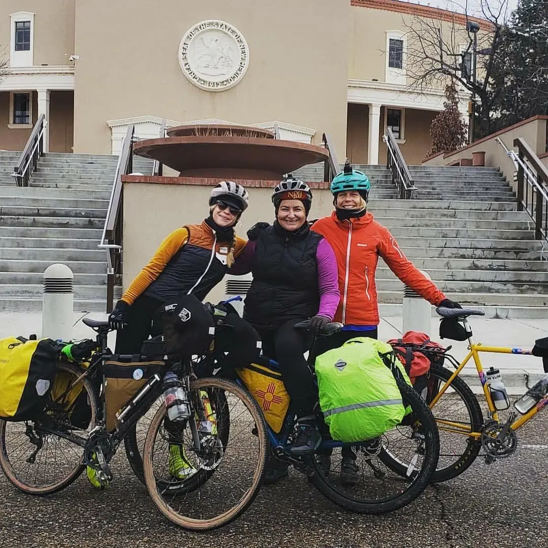Three cyclists in helmets and winter gear smile for a photo in front of the New Mexico State Capitol, each with touring bicycles loaded with brightly colored panniers