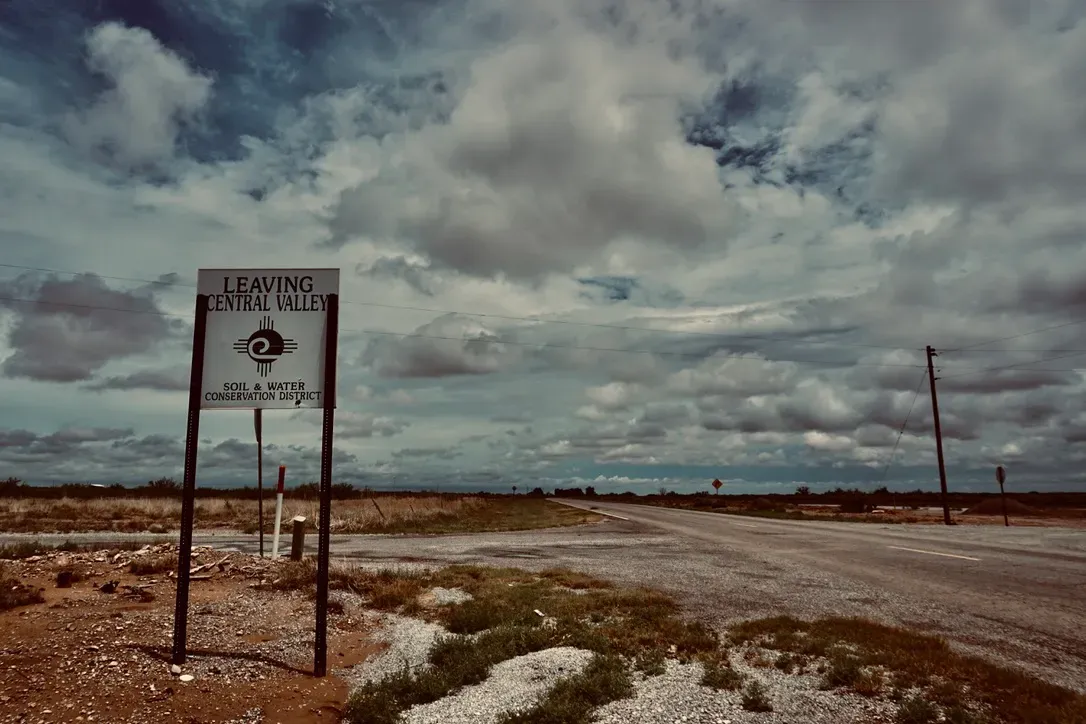 Roadside sign reading “Leaving Central Valley Soil & Water Conservation District” stands beside an empty rural highway under a dramatic, cloudy sky
