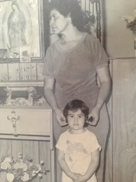 Black-and-white photo of a woman standing behind a young girl, gently holding her ears, with a home altar featuring a crucifix, flowers, and an image of Our Lady of Guadalupe in the background