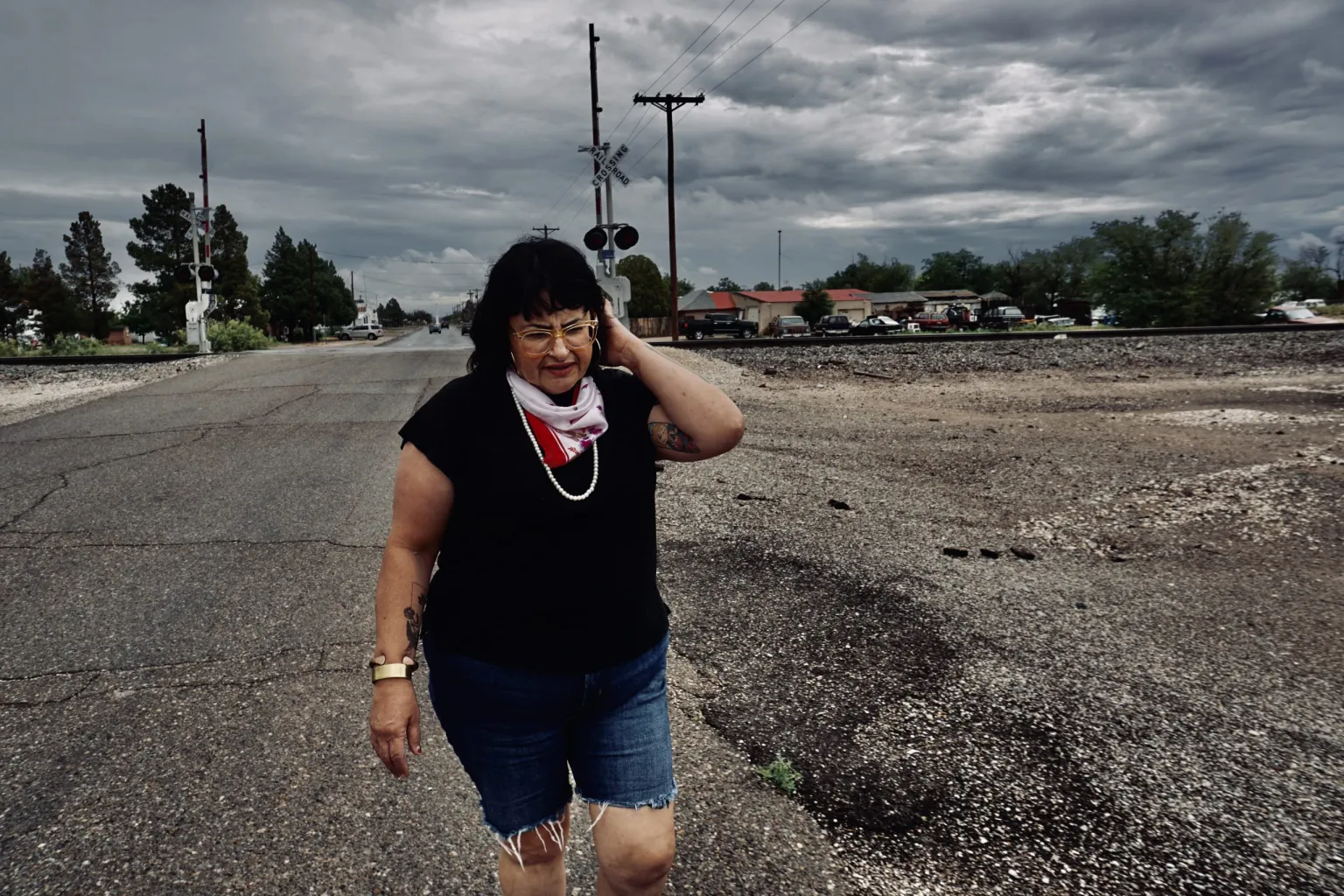 A woman wearing a black shirt, denim shorts, and a red and white scarf walks along a road near railroad tracks under a cloudy, stormy sky