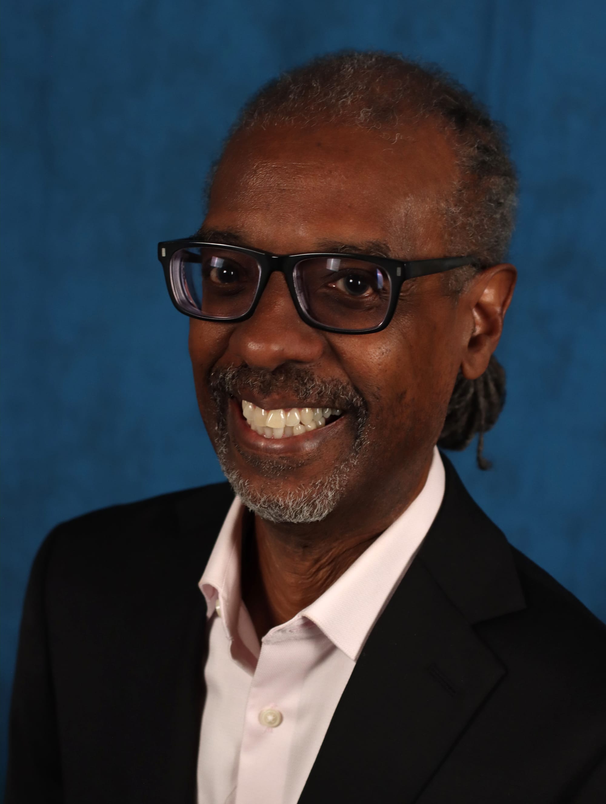 Gene Grant smiles while wearing glasses, a dark blazer and a light pink shirt against a blue backdrop.
