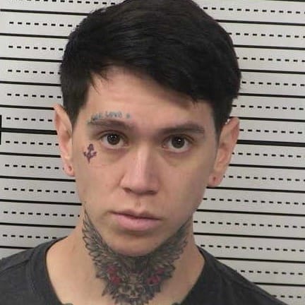 Booking photo of Isaac James Brooks, a 27-year-old man with short dark hair and neck and face tattoos, taken at the Doña Ana County Detention Center.