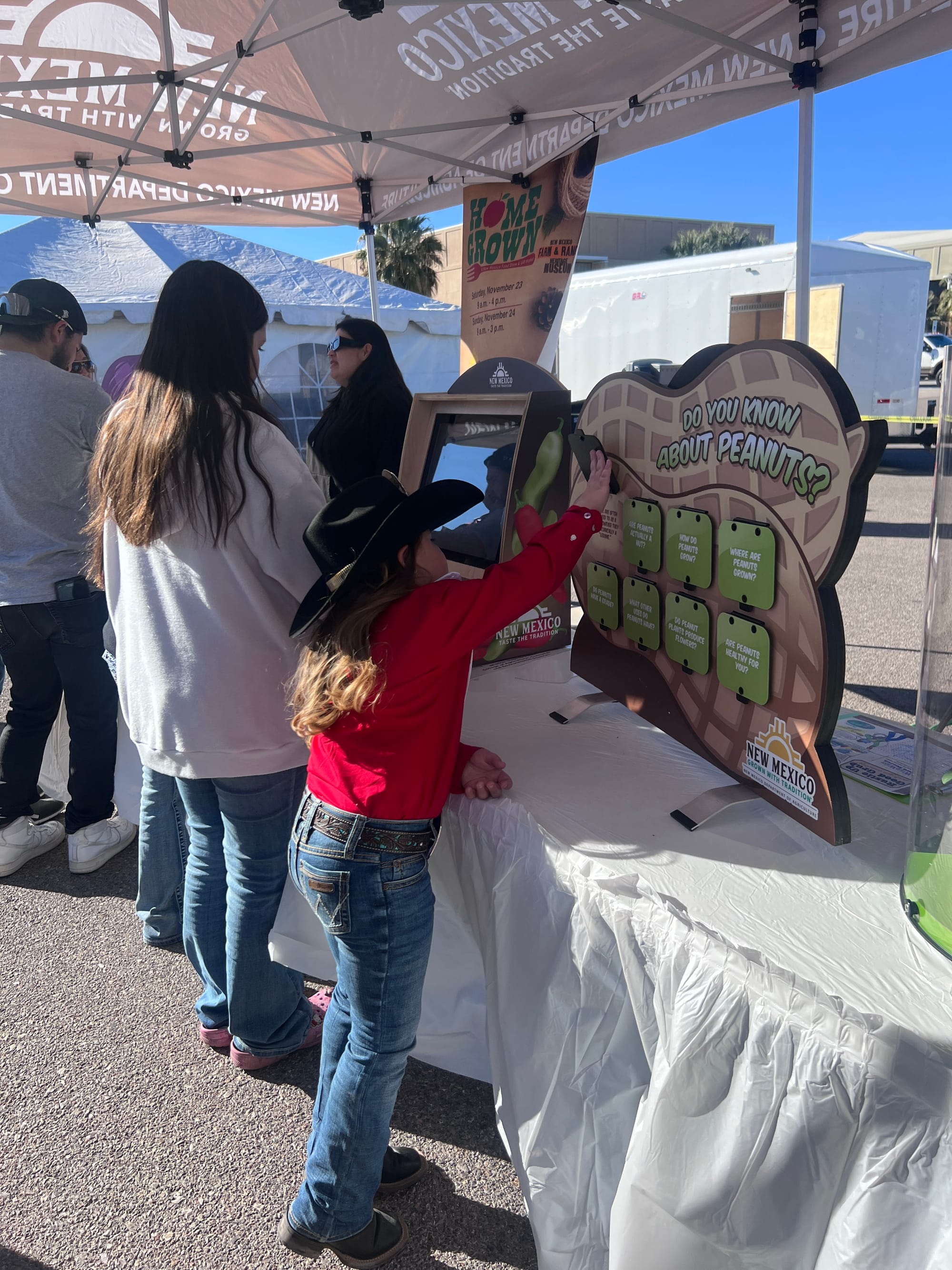 A young girl wearing a black cowboy hat and red shirt reaches toward an interactive display about peanuts under a New Mexico Department of Agriculture tent during Ag Day at New Mexico State University.