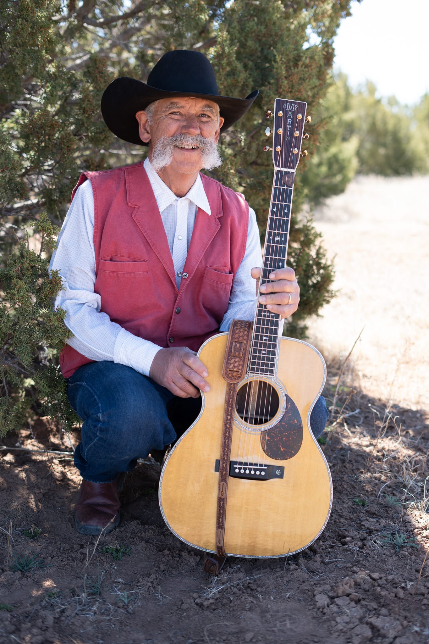 Singer-songwriter and rancher Randy Huston stands outdoors wearing a black cowboy hat and red vest, smiling while holding his Martin guitar near juniper trees in rural New Mexico.