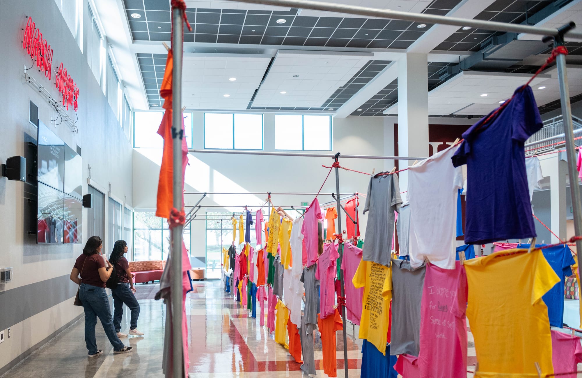 Two visitors stand beside rows of colorful T-shirts hanging on lines inside NMSU’s Corbett Center during a past Clothesline Project exhibit.