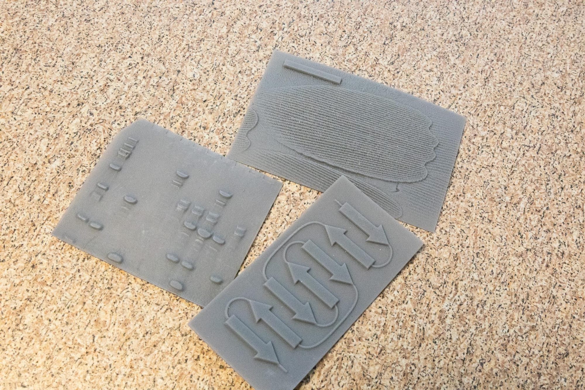 Three tactile 3D-printed gray sheets with raised molecular and structural patterns rest on a tabletop, designed to help visually impaired students interpret complex scientific data.