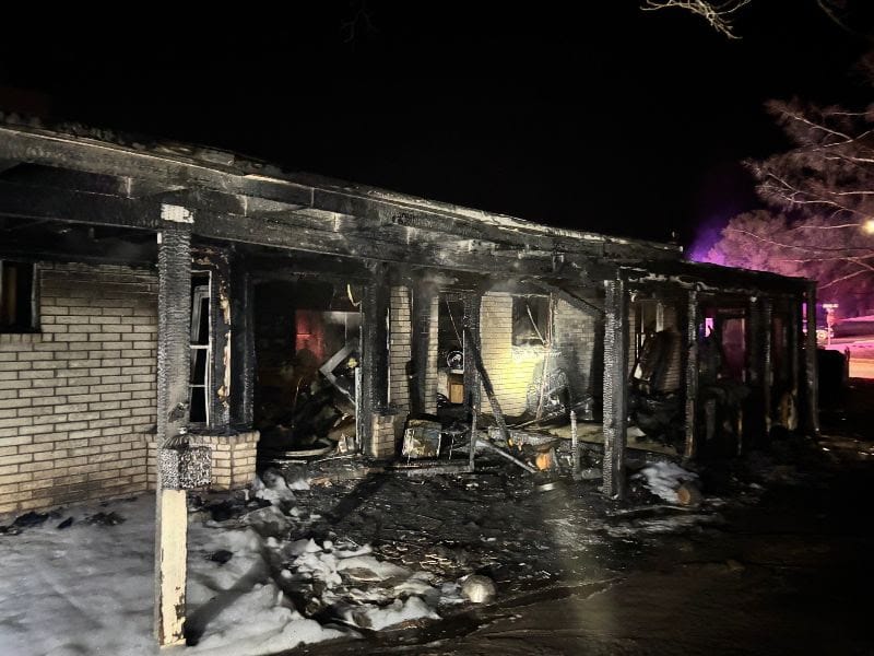 A one-story home on Enchanted Drive is heavily damaged after a fire, with charred porch supports, burned debris and foam on the ground as emergency lights glow in the background.