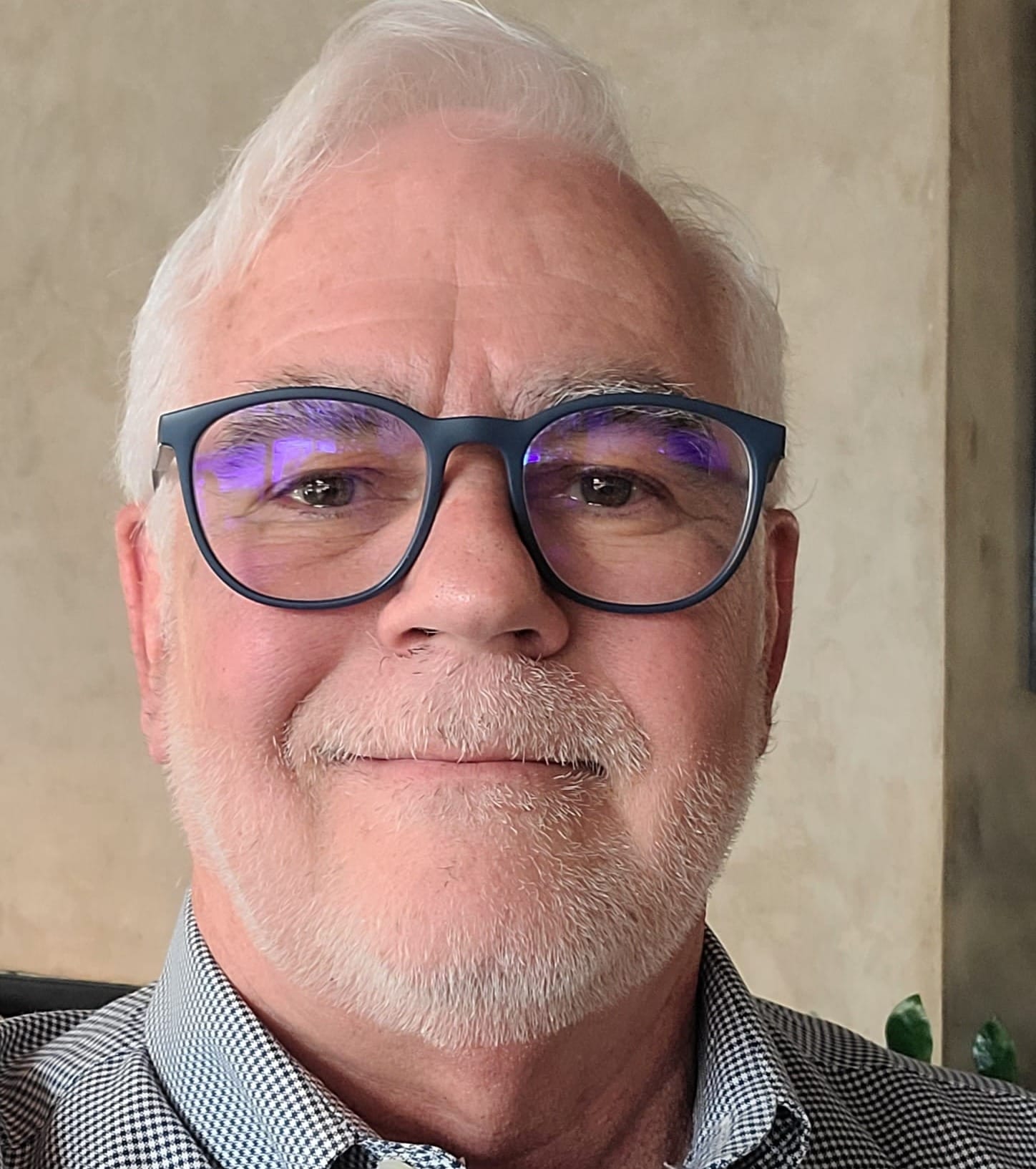Michael Marcotte, with white hair, a trimmed white beard and blue-rimmed glasses smiles slightly at the camera while wearing a checkered shirt indoors.