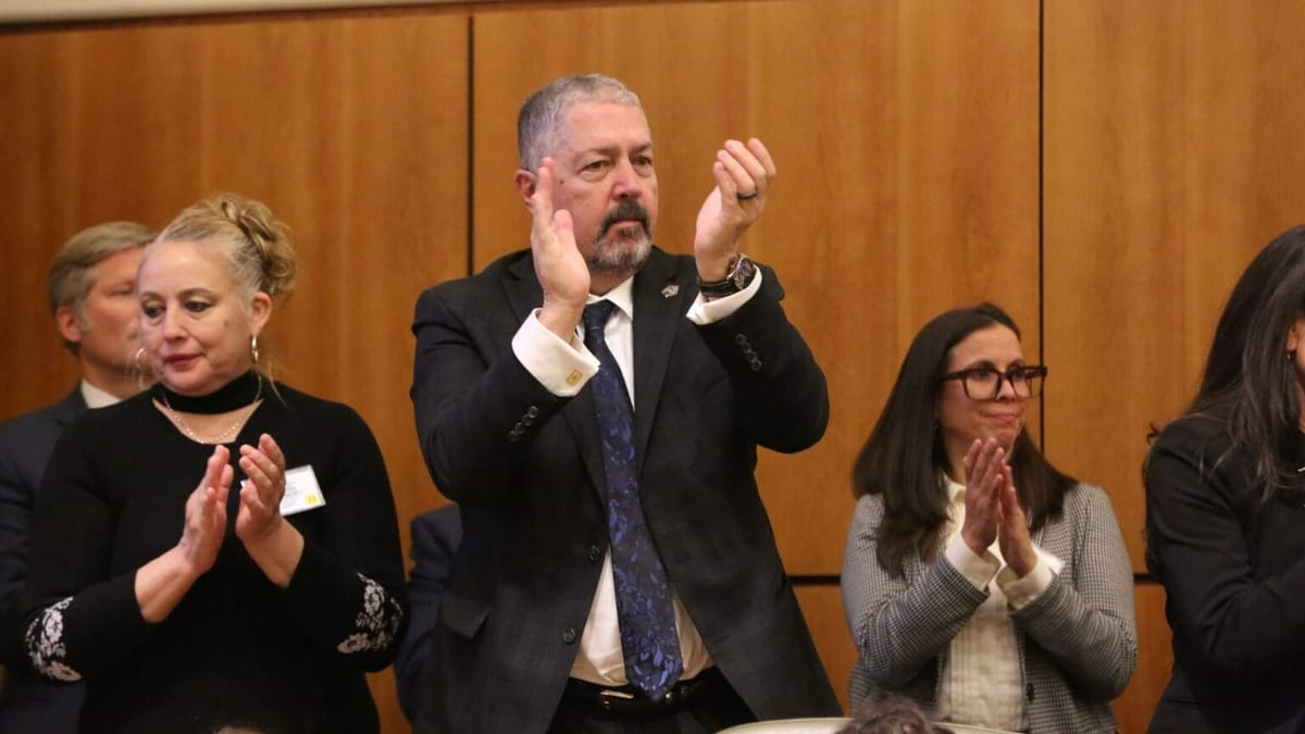 A group of people stand and applaud during an indoor event, with Sam Bregman, in a dark suit and blue tie, clapping at center and others clapping beside him against a wood-paneled background.