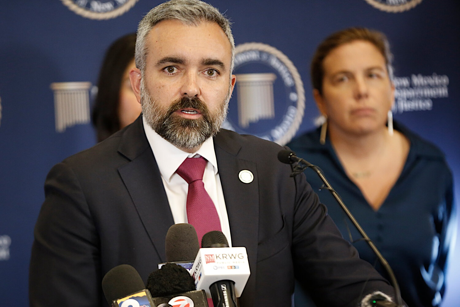 New Mexico Attorney General Raúl Torrez speaks at a press conference behind a cluster of microphones, wearing a dark suit and red tie, with a blurred backdrop featuring the Department of Justice seal and staff members standing behind him.