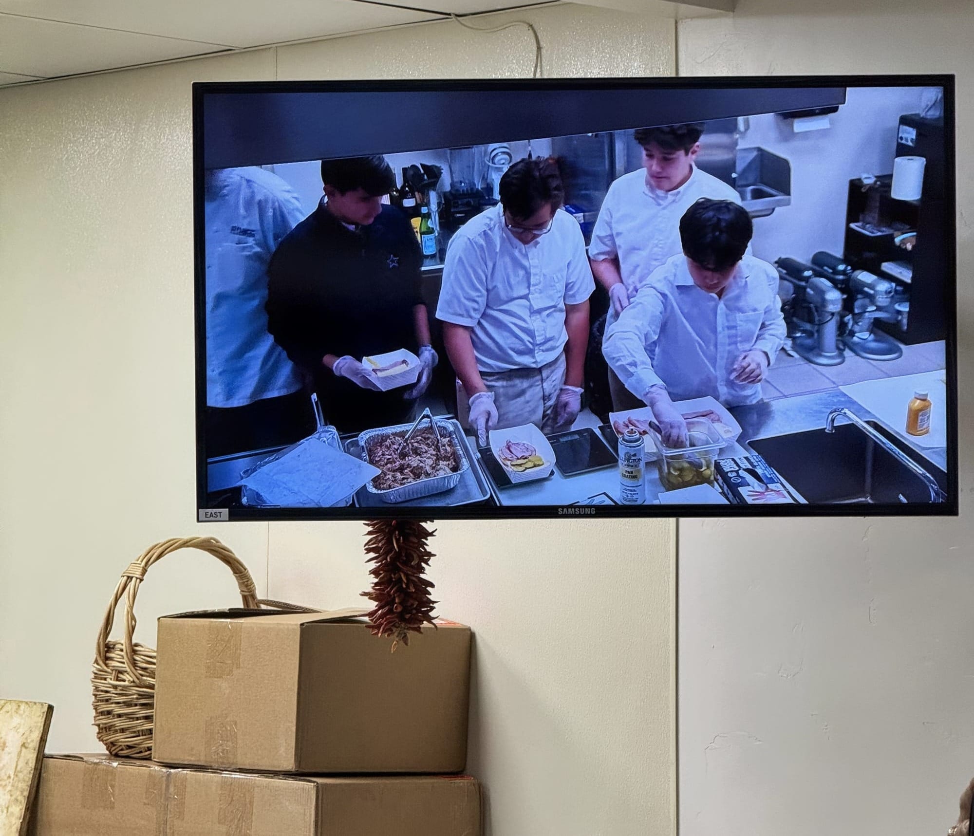 A wall-mounted screen shows middle school students assembling Cubano paninis in a commercial kitchen during a culinary arts demonstration at DACC.