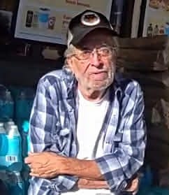 A 68-year-old man wearing a dark baseball cap, glasses, and a blue plaid shirt sits indoors with his arms crossed. He has gray facial hair and a serious expression.