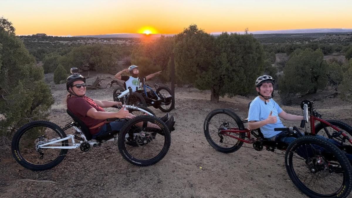 Three participants ride adaptive recumbent bikes on a desert trail at sunset during a group outdoor recreation activity supported by New Mexico’s Outdoor Equity Fund.