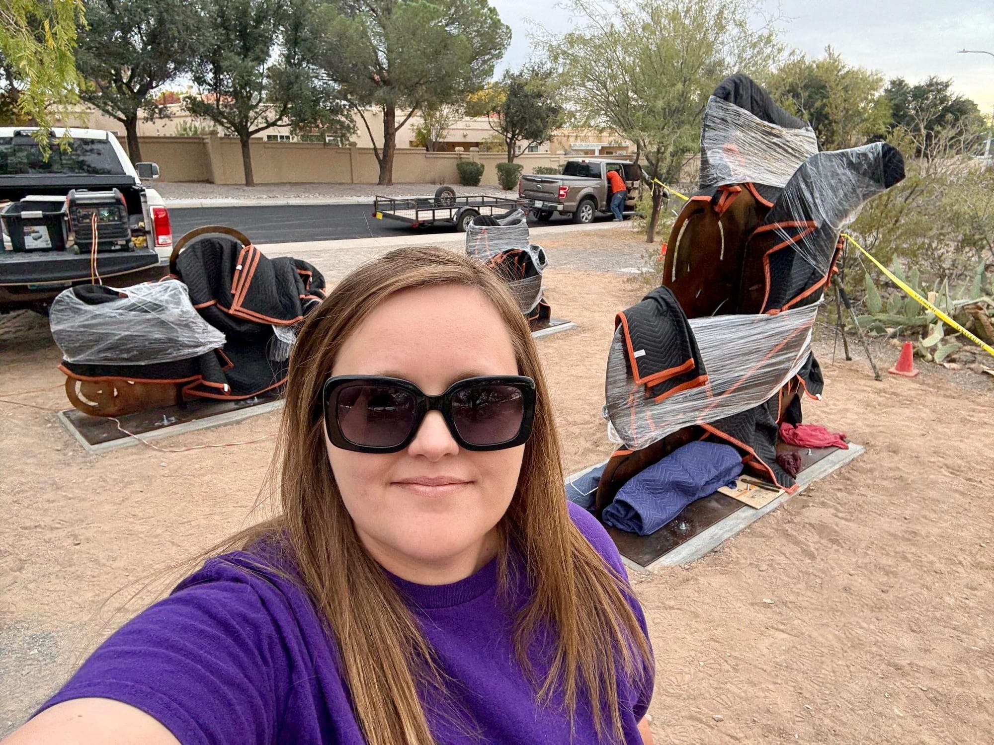 Kayla Blundell takes a selfie at a Las Cruces park while metal sculpture pieces wrapped in protective padding sit on concrete bases during installation, with trucks and tools visible nearby.