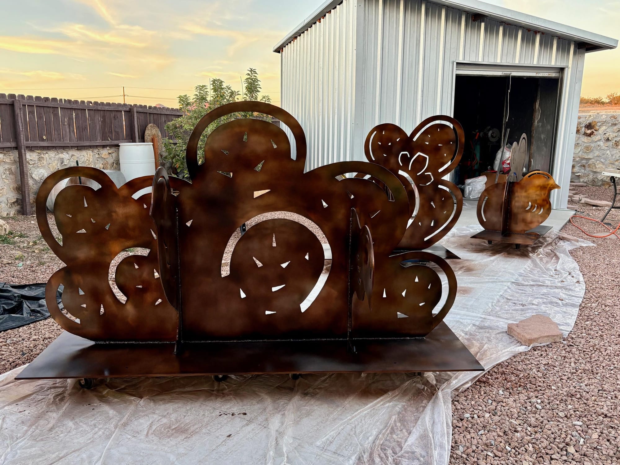 Metal sculpture panels shaped like desert plants rest on a base outside a workshop, with additional pieces nearby as the public art installation is prepared.
