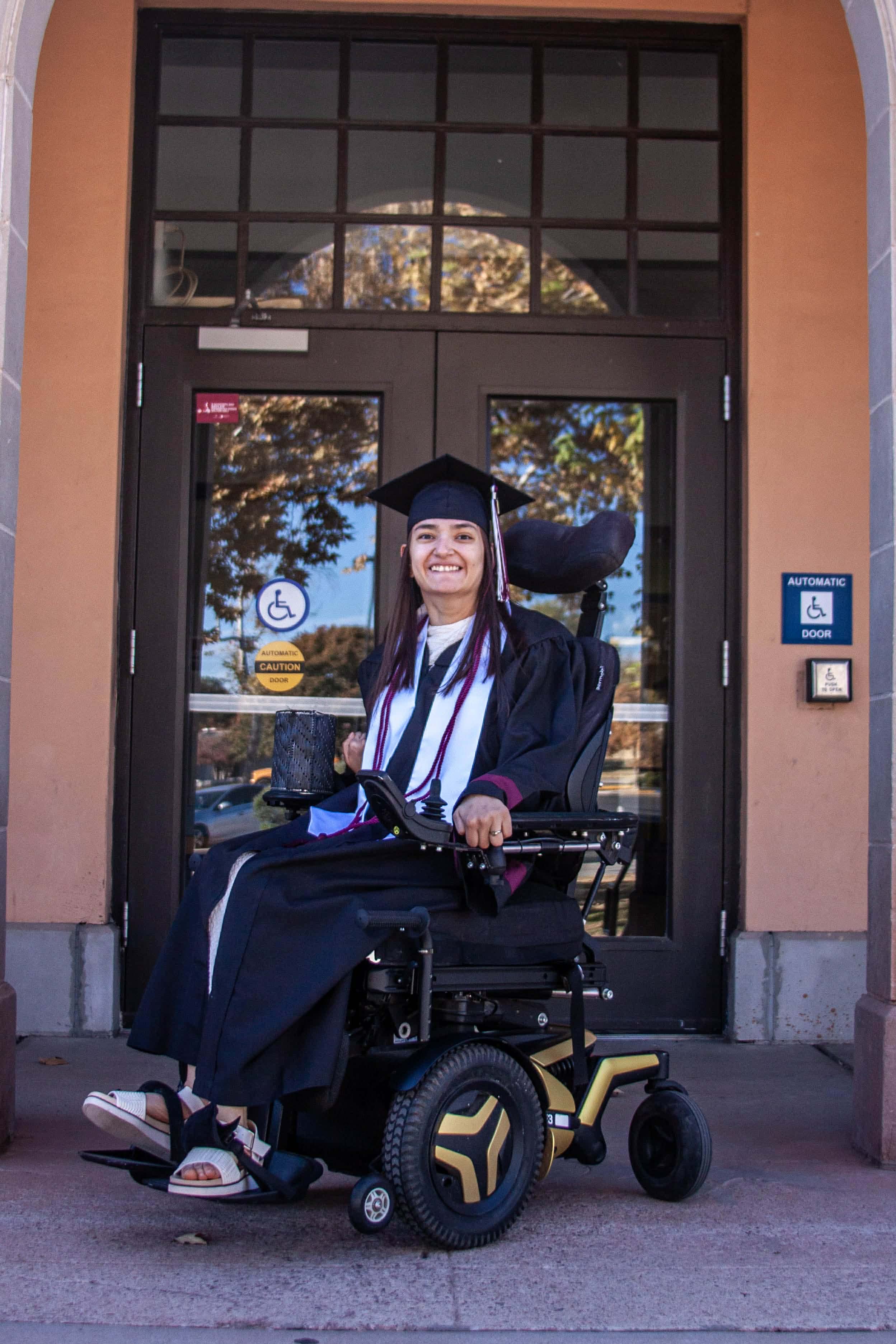 Arcelia Mendoza, wearing a graduation cap and gown, smiles while seated in a powered wheelchair outside an NMSU campus building.