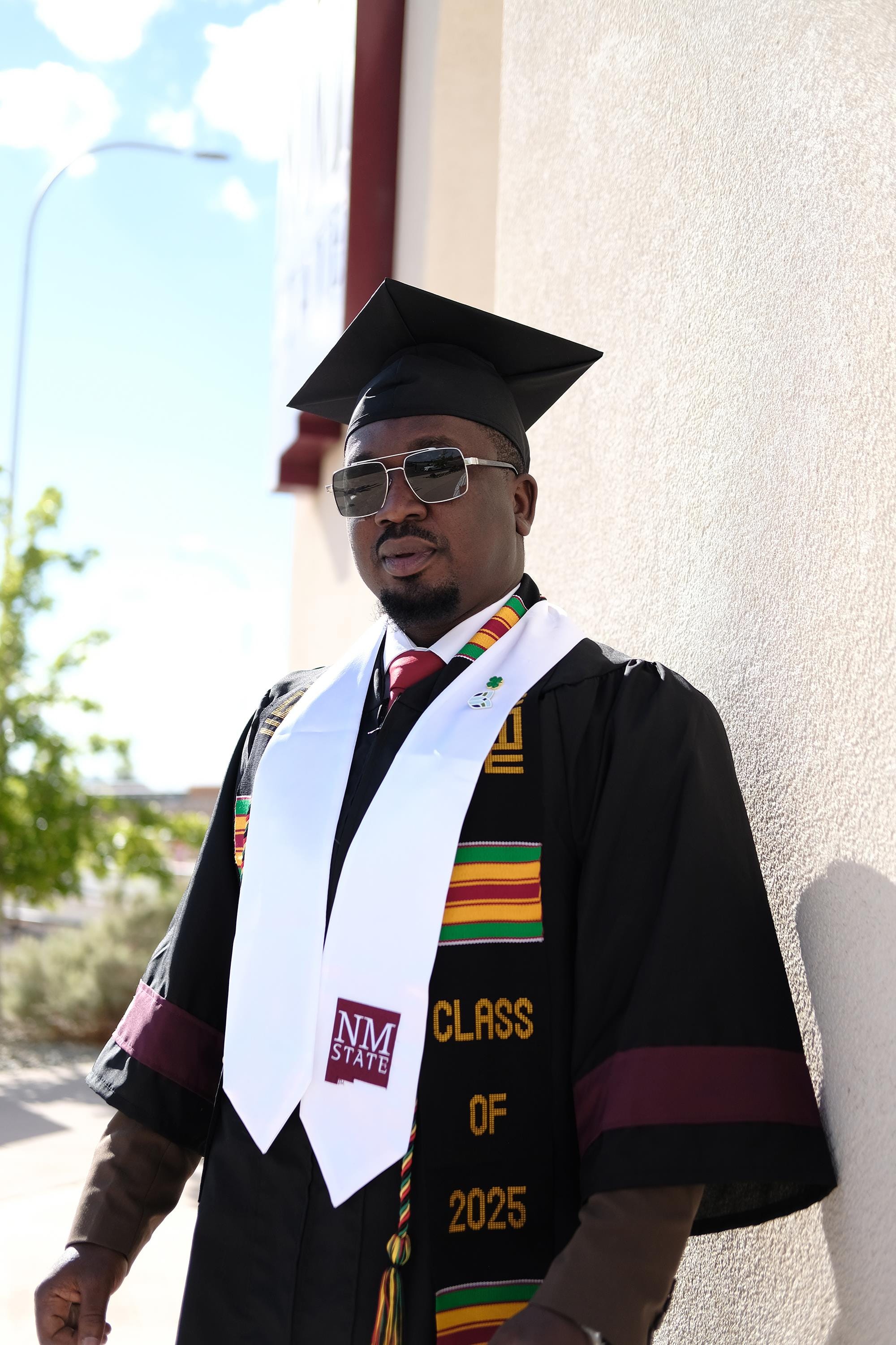 Kazeem Abdulrazaq, wearing a cap and gown with an NMSU Class of 2025 stole, stands outdoors against a building wall, wearing sunglasses and ceremonial honor cords.