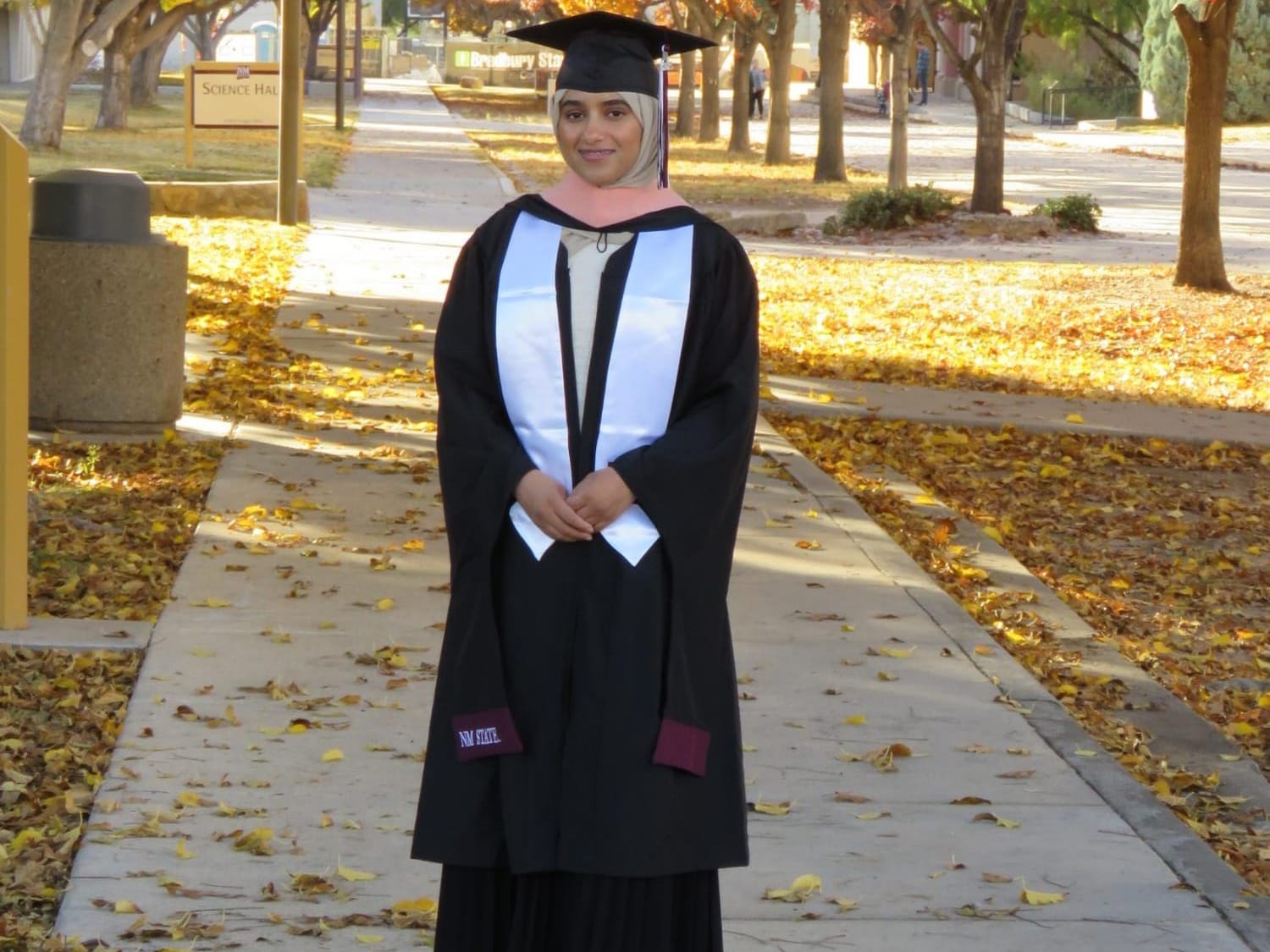 Aya Abdaljalil stands on the NMSU campus wearing a graduation cap and gown, posing on a tree-lined walkway covered with autumn leaves.