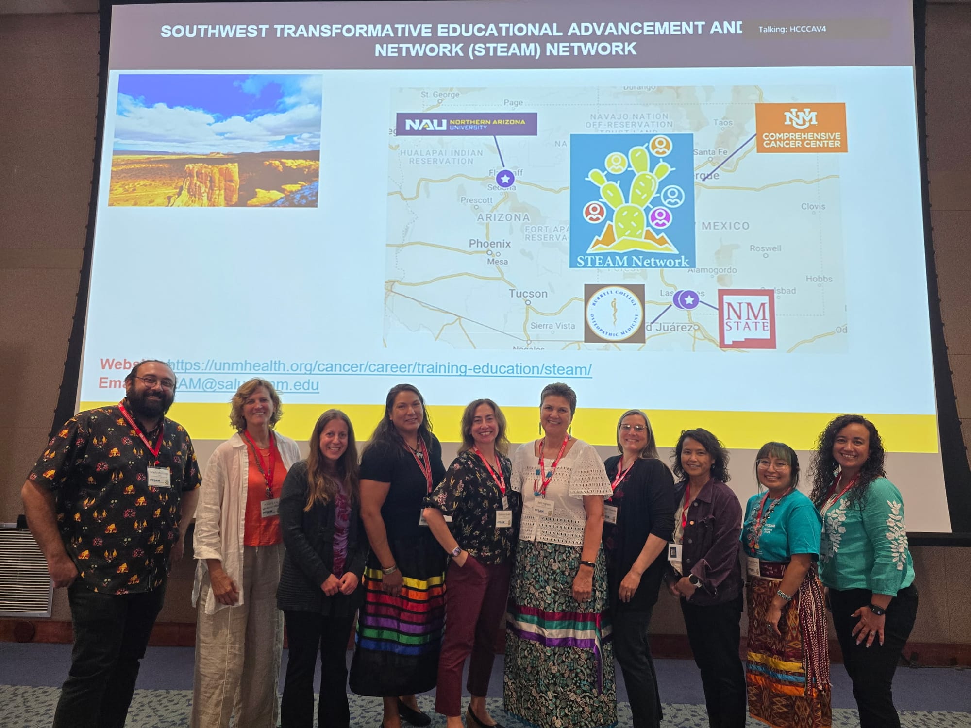 A group of researchers stand together at a conference, posing in front of a projected screen displaying a map of the Southwest and logos for NMSU, UNM and the STEAM Network.