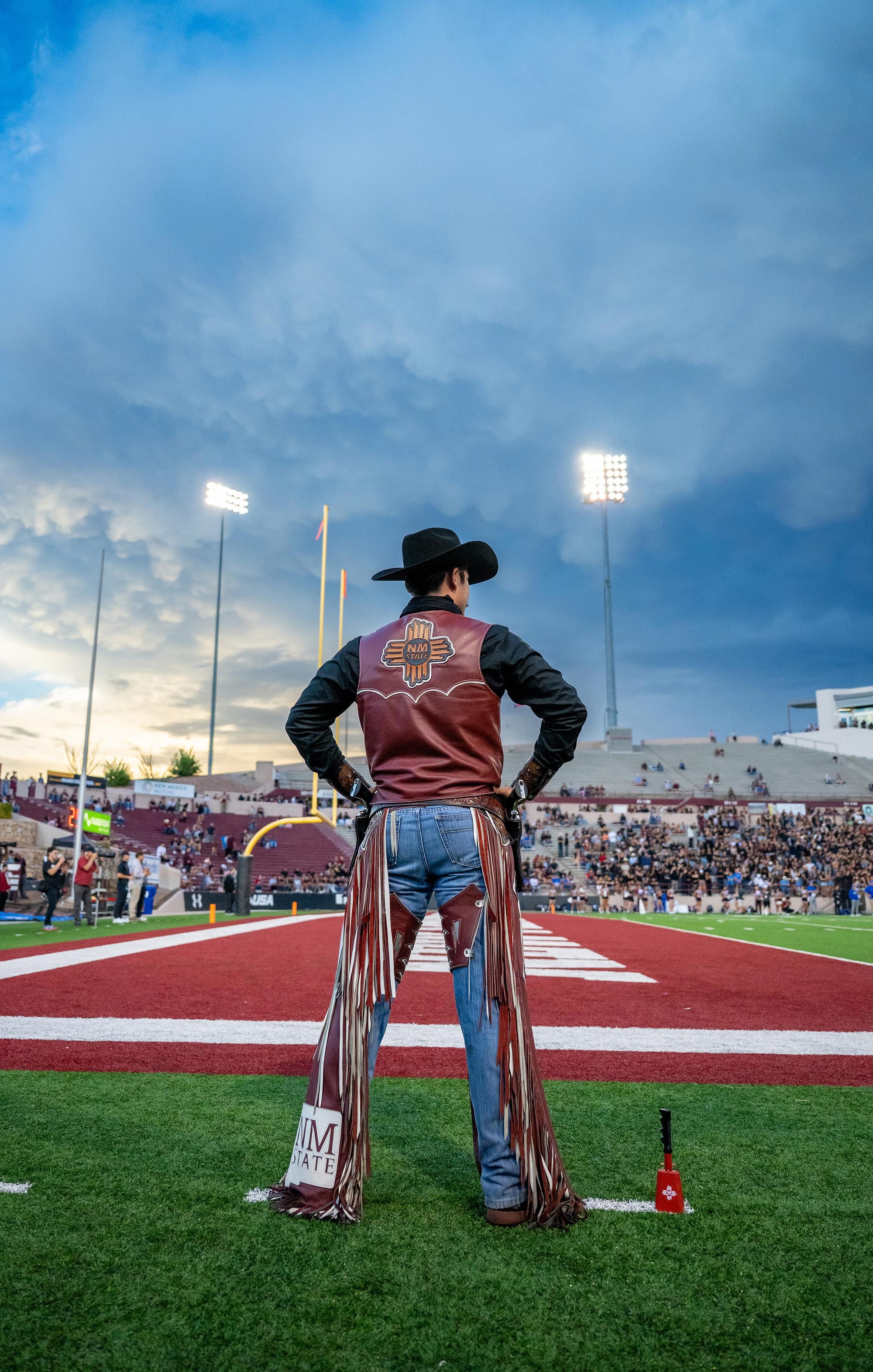 Pistol Pete stands on the field at Aggie Memorial Stadium during a football game, seen from behind facing the crowd under stadium lights.