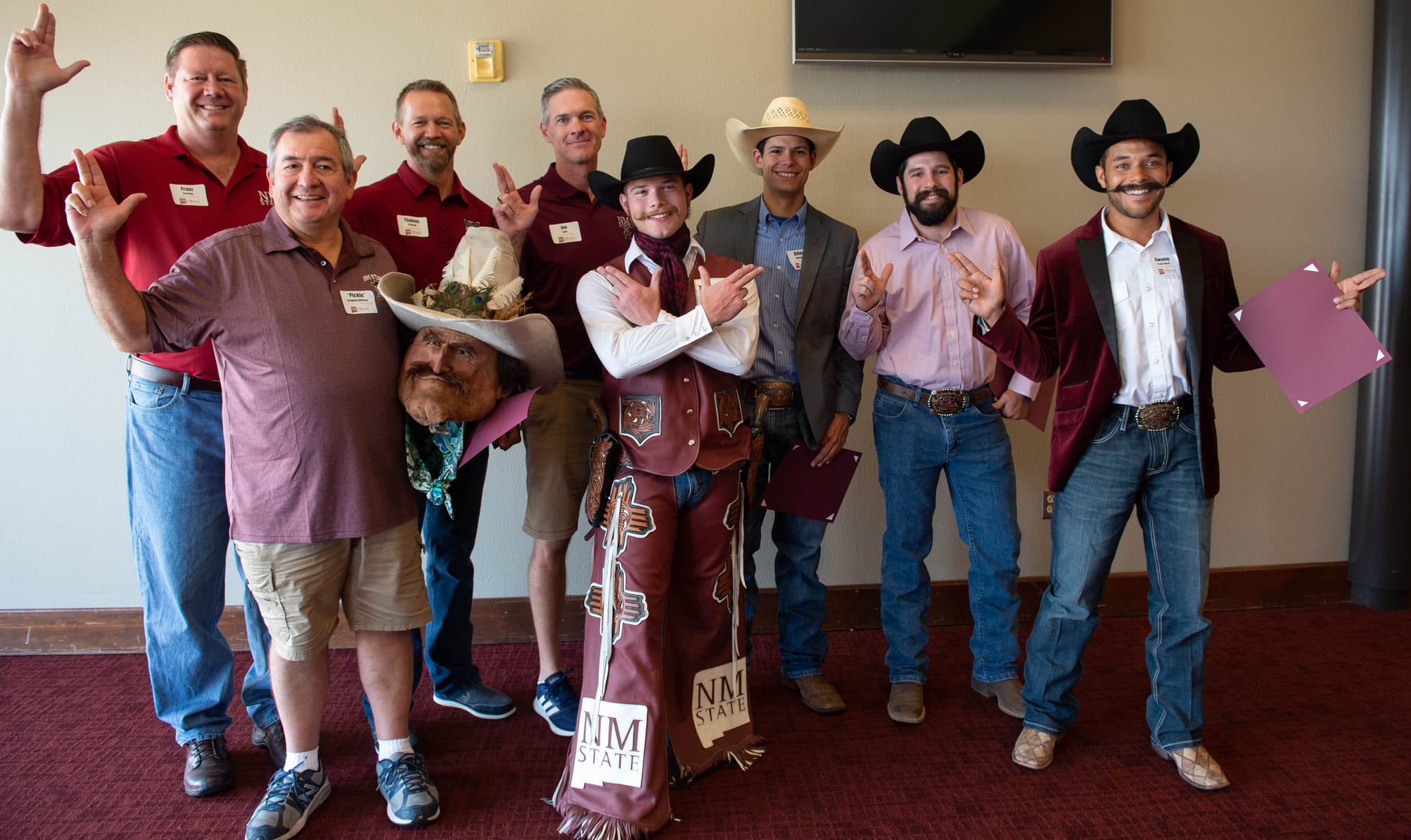 Past Pistol Pete performers pose indoors with NMSU staff and alumni, wearing Aggie apparel and cowboy hats, some holding certificates and making hand signs.