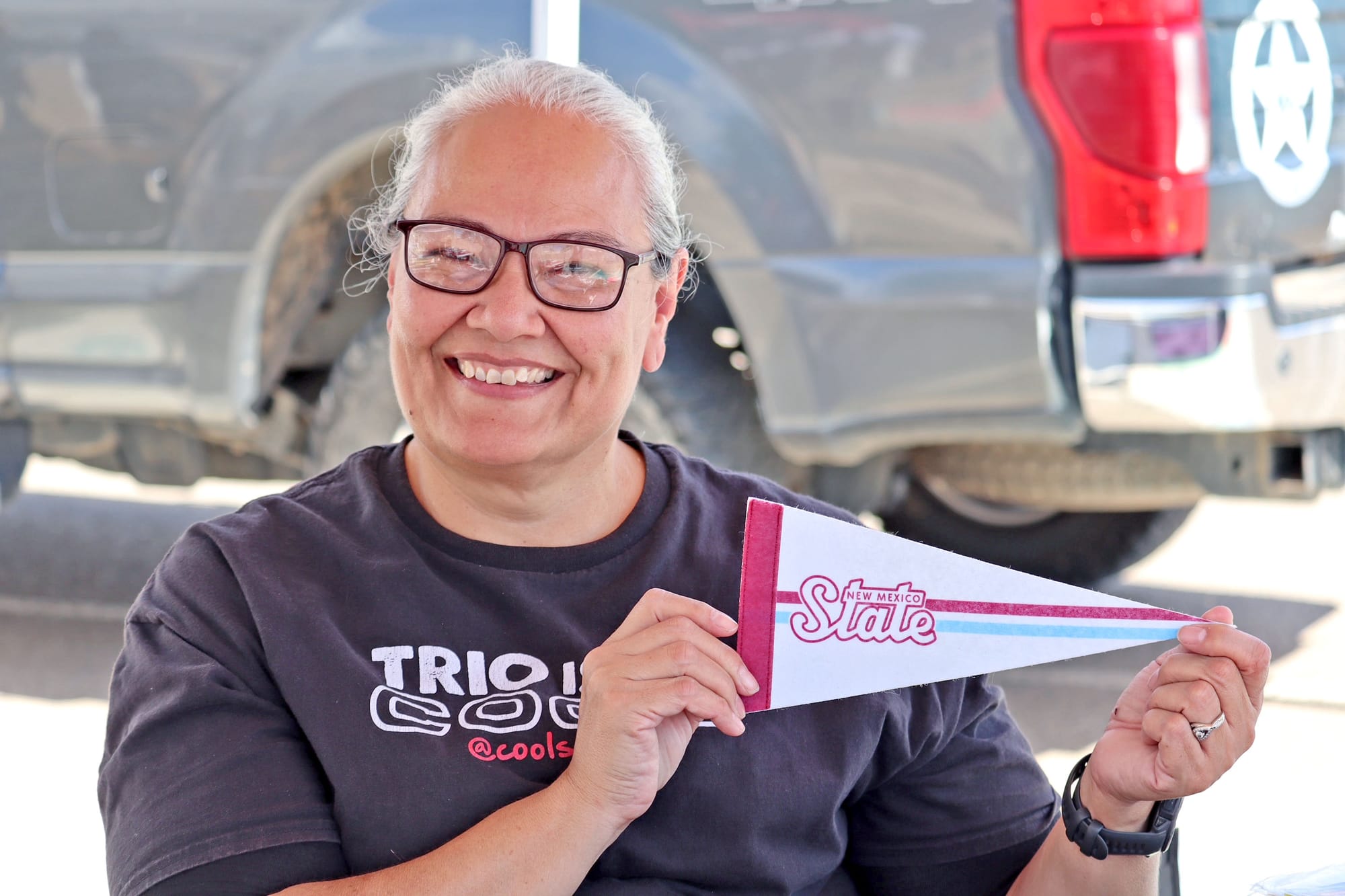 Lourdes Ambriz, wearing a TRIO program T-shirt, smiles while holding a New Mexico State University pennant during an outreach event focused on college readiness.