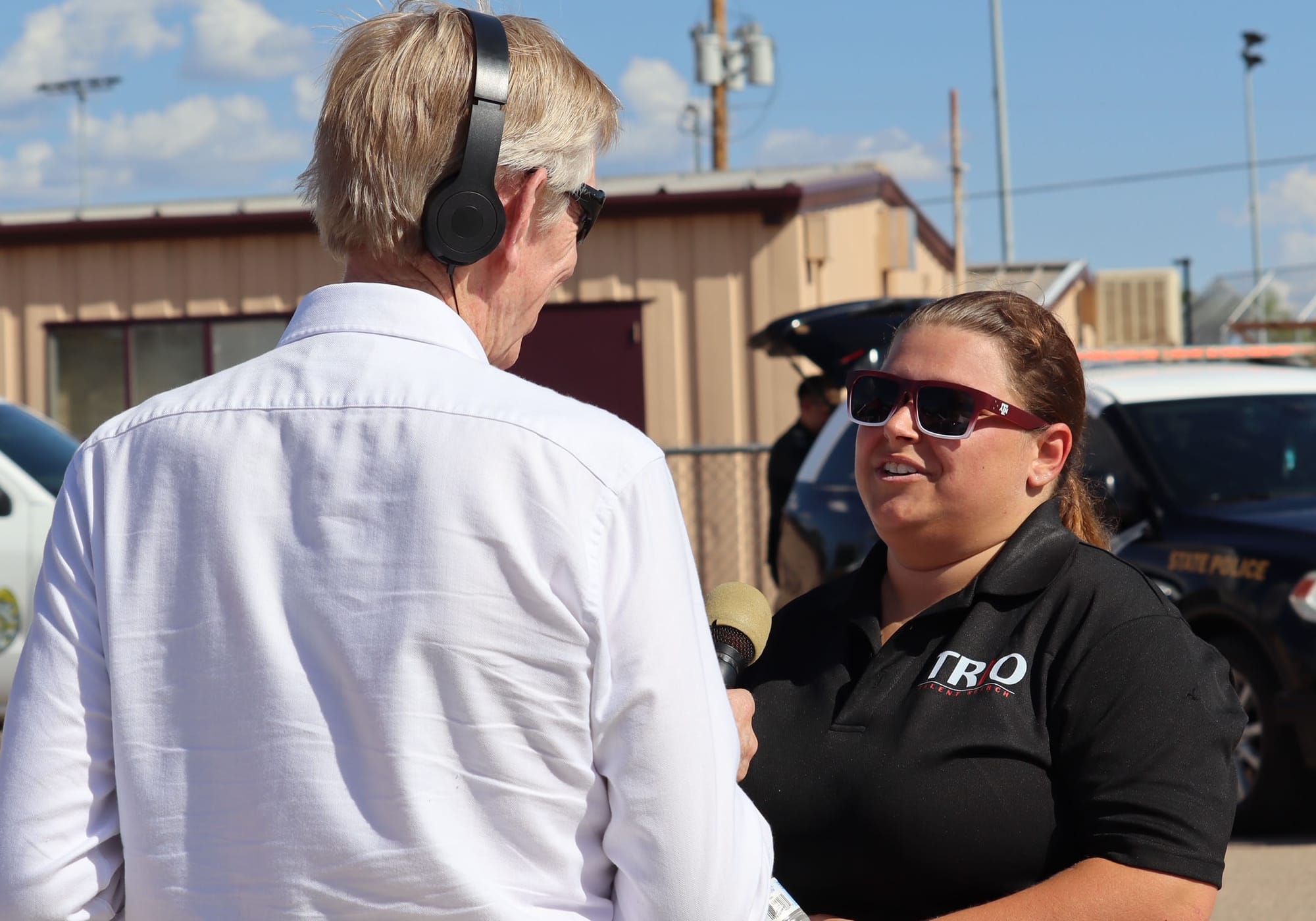 A TRIO staff member speaks with a reporter holding a microphone during an outdoor college outreach event, with vehicles and buildings visible in the background.