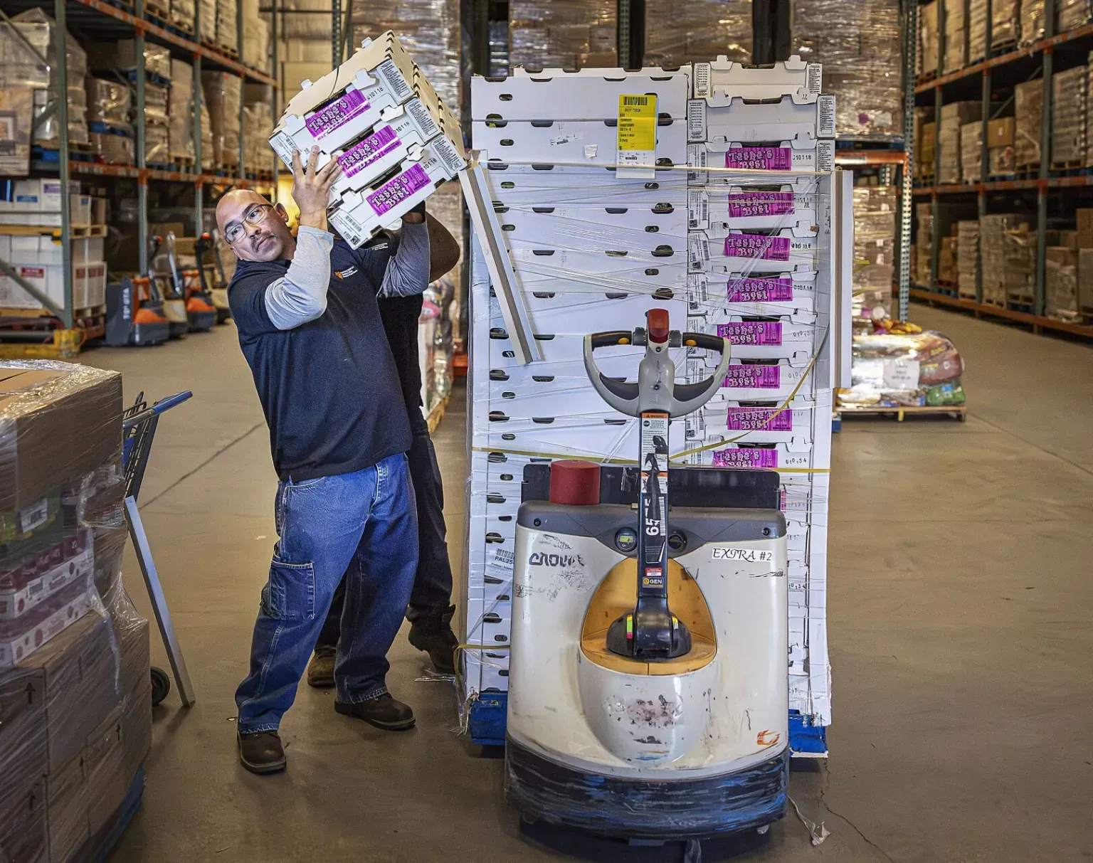 A worker lifts stacked boxes of produce onto his shoulder inside a large warehouse, standing beside a pallet jack loaded with shrink-wrapped cartons and surrounded by shelving filled with food.