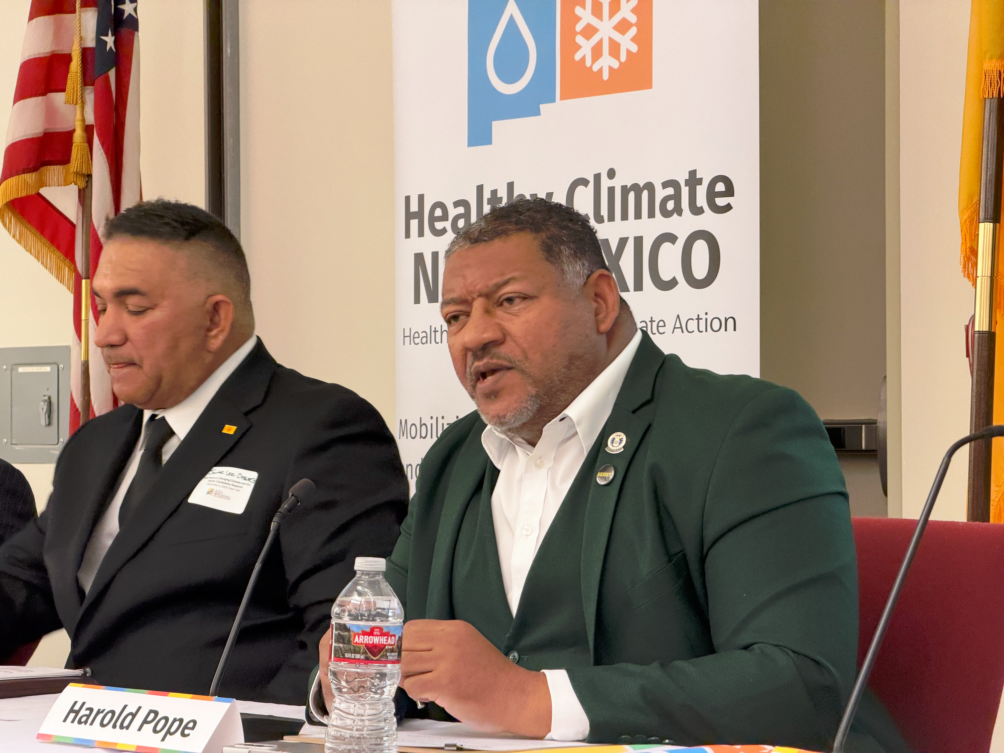 Harold Pope speaks during a Healthy Climate New Mexico town hall panel, seated at a table with microphones, name placards and the organization’s banner visible behind him.