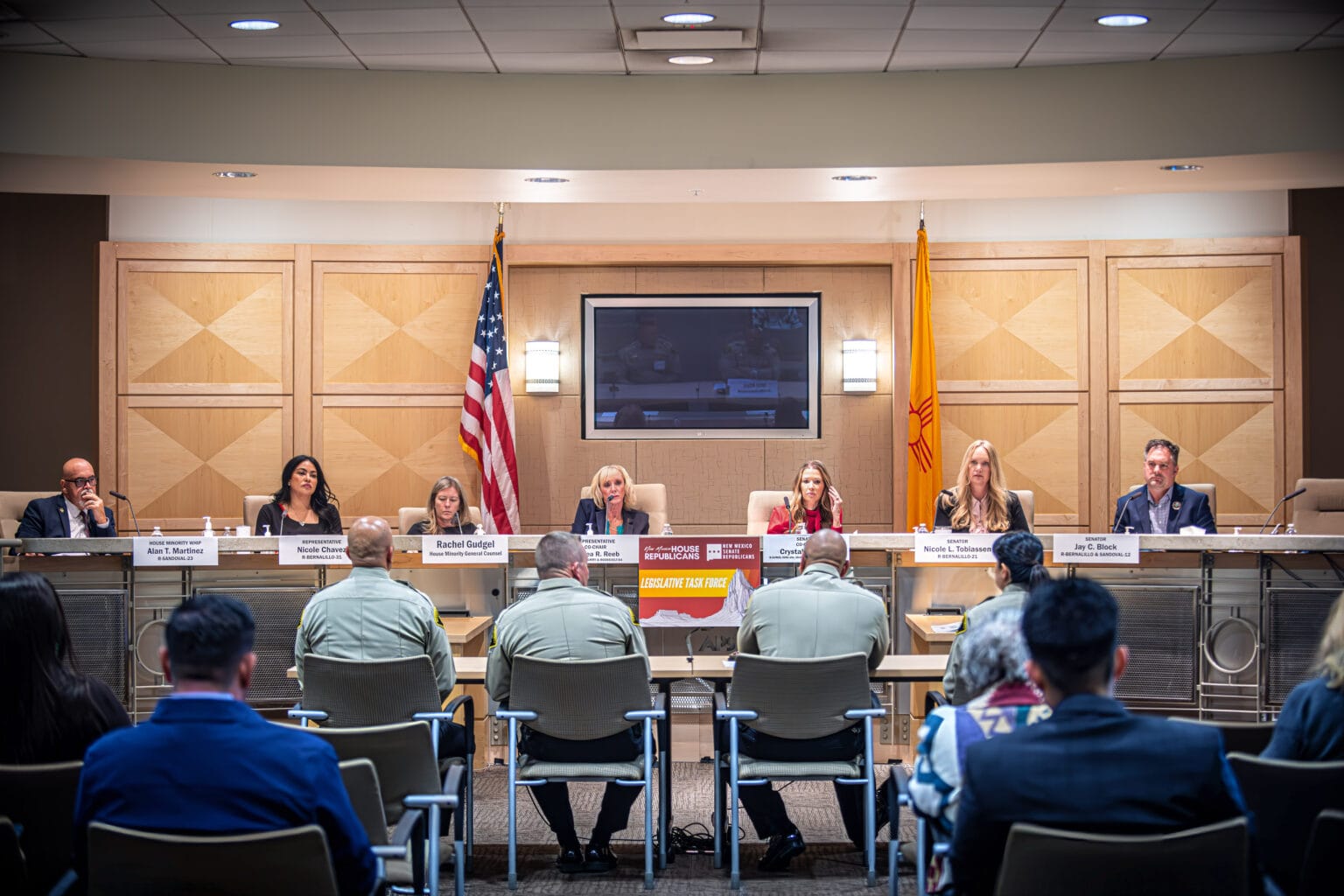 A panel of New Mexico lawmakers sits at a dais during a legislative task force meeting, with law enforcement officers and members of the public seated in the audience beneath U.S. and New Mexico flags.