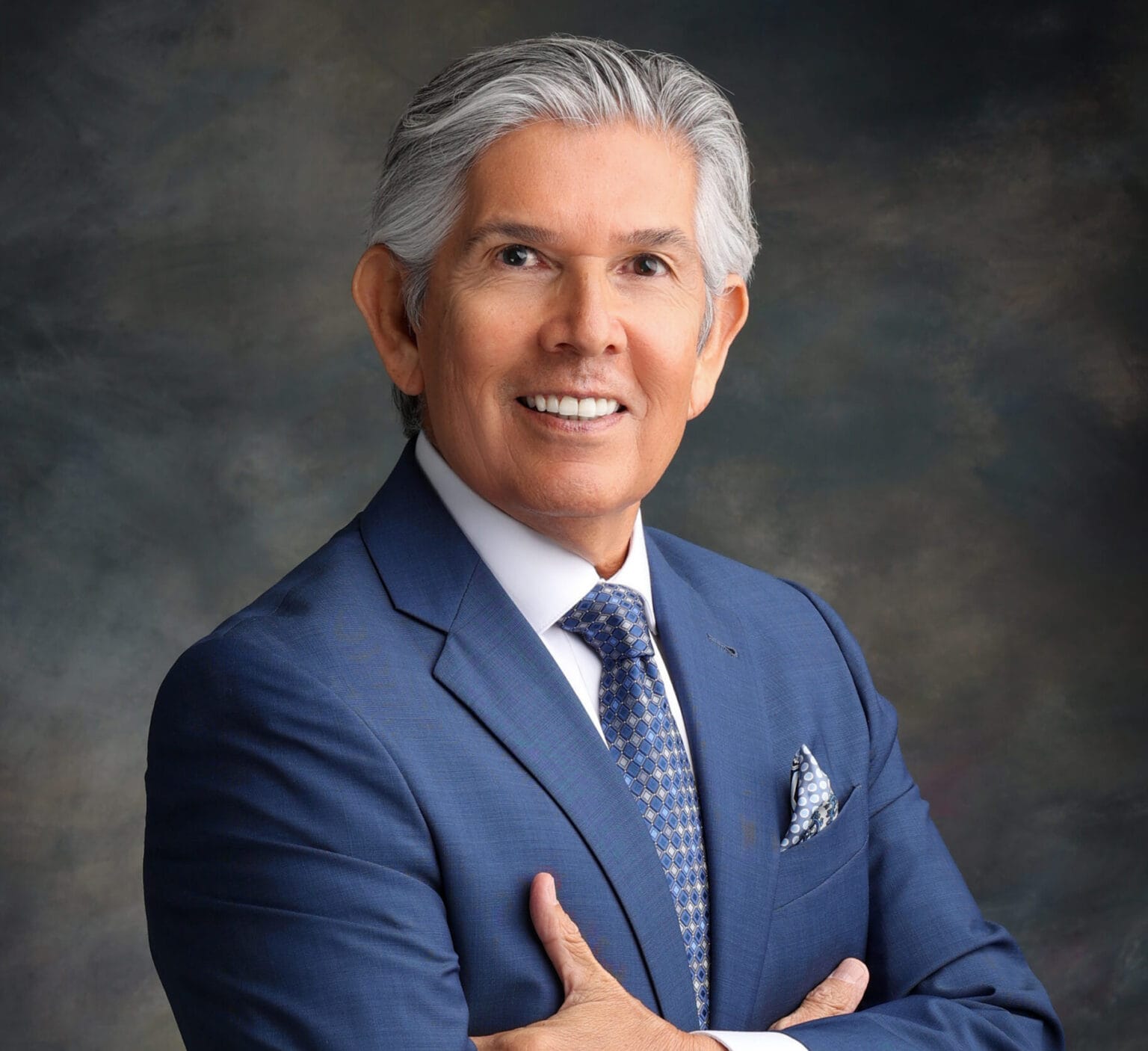 Duke Martinez, with gray hair wearing a blue suit and patterned tie, poses for a studio portrait with his arms crossed.