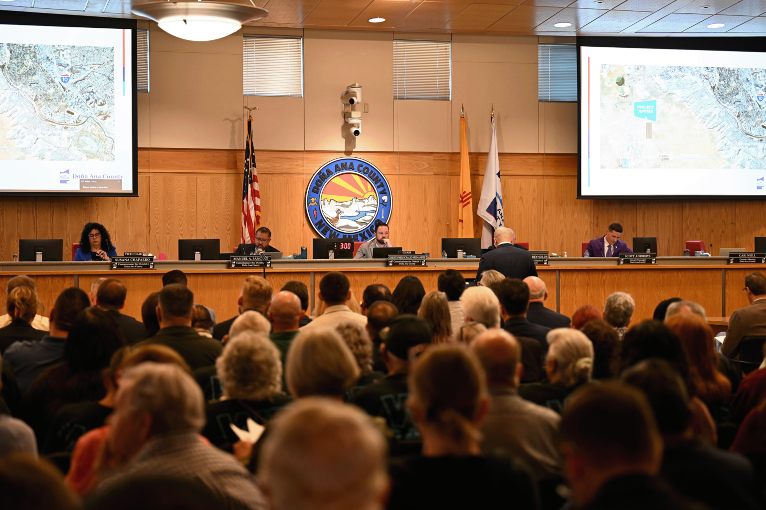 Residents fill the Doña Ana County Commission chambers as officials discuss Project Jupiter during a public meeting, with maps displayed on screens at the front of the room.