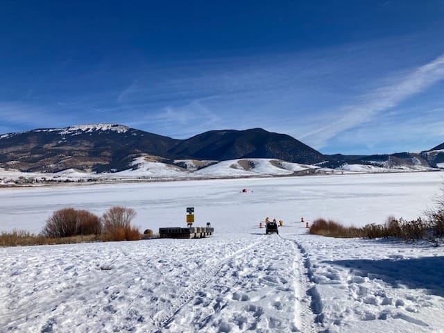 Snow-covered shoreline and boat ramp at a frozen New Mexico state park lake, with mountains rising in the background under a clear blue winter sky.