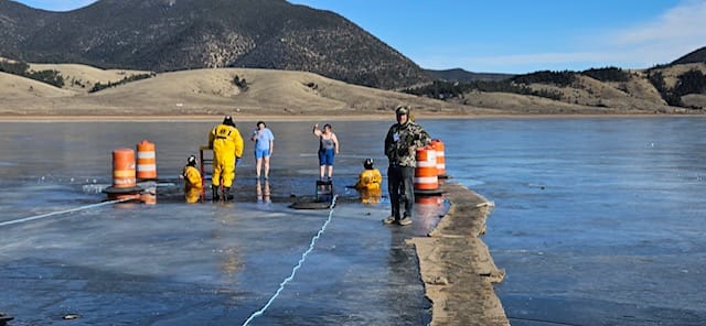 Participants gather at a marked opening in a frozen lake during a polar bear plunge at a New Mexico state park, with safety personnel and snow-covered hills in the background.