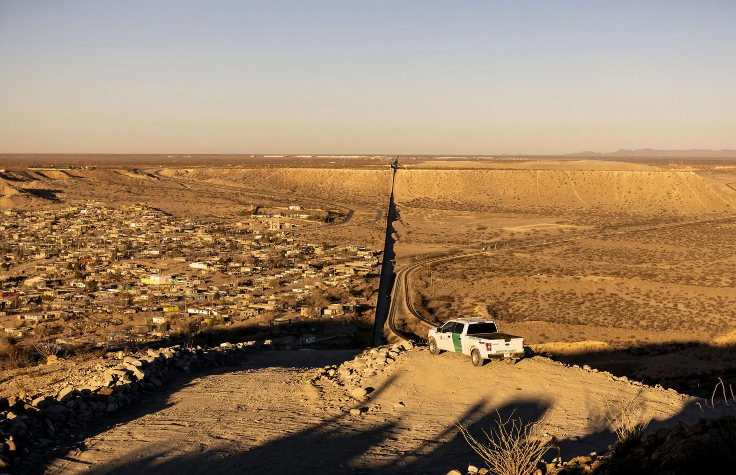 The U.S.-Mexico border wall cuts across a desert landscape near a densely built border community, with a Border Patrol vehicle parked on a dirt road overlooking the divide.