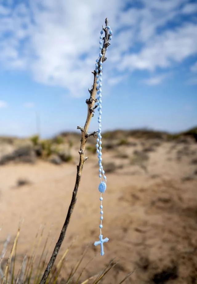 A blue rosary hangs from a dry branch in a sandy desert landscape.