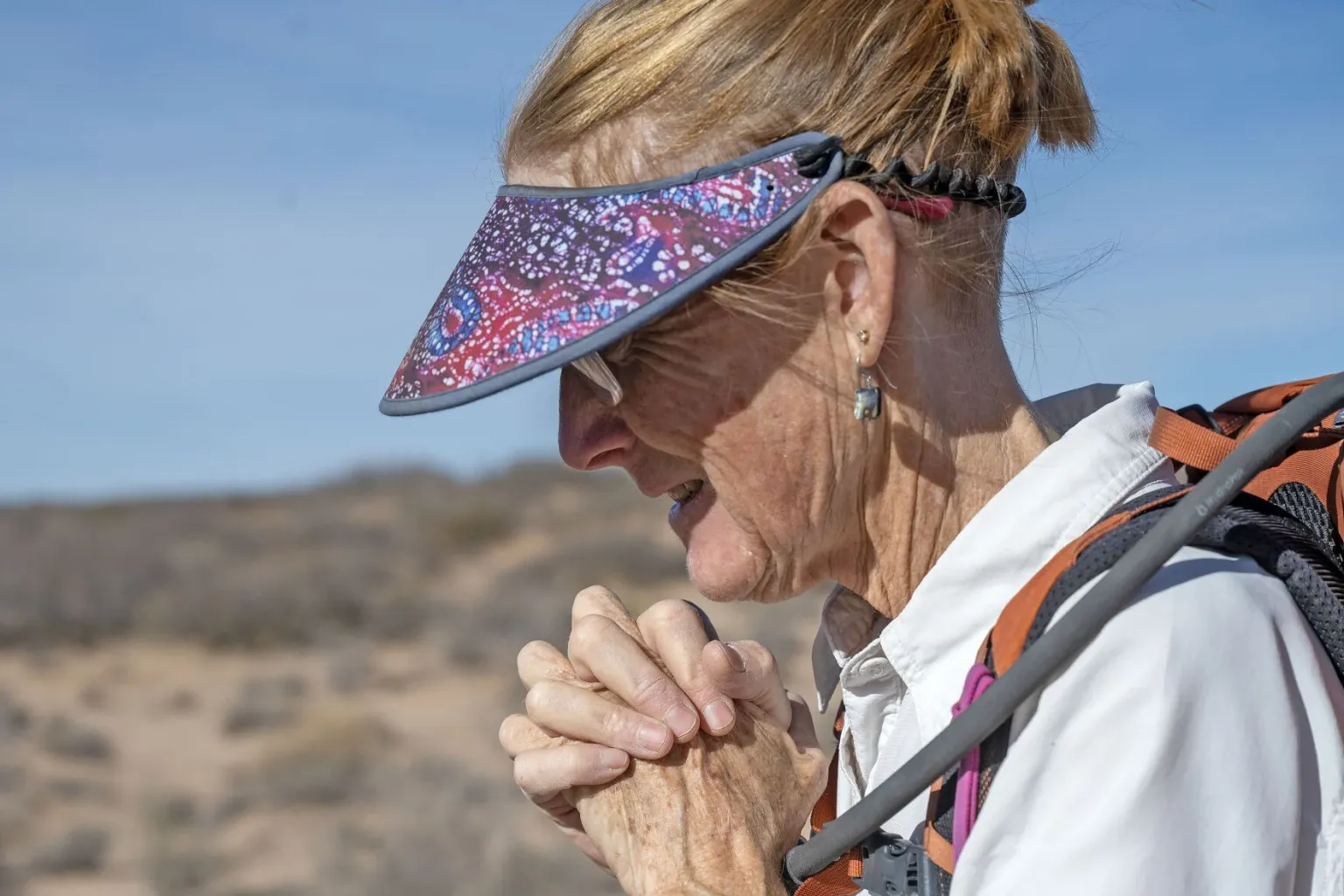 A woman wearing a visor and backpack stands in a desert landscape with her hands clasped near her face, appearing reflective or concerned.